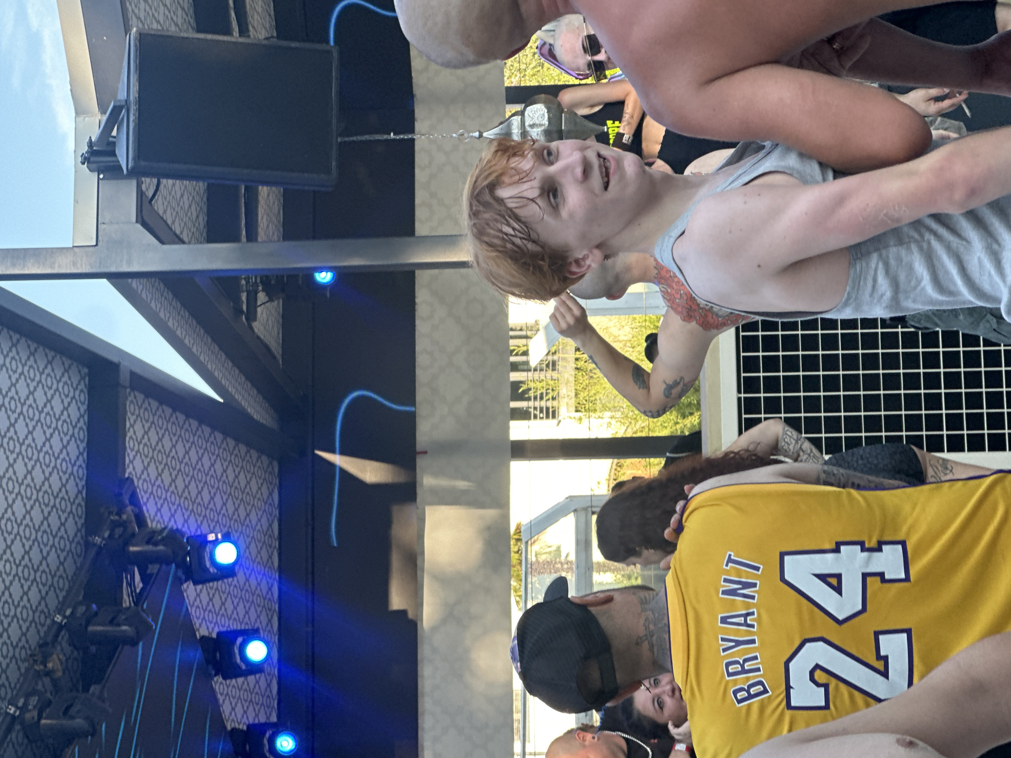 The image shows a group of people gathered outdoors, possibly at an event or concert. A young person with light-colored hair and a gray tank top is in the foreground, smiling and looking to the right. To the left, a person wearing a yellow Kobe Bryant #24 jersey is visible from the back. In the background, there is a stage area with blue lights and a patterned ceiling. The setting appears to be a public gathering space with an open-air roof structure. There are no indications that this image is located in Borgonato, Italy.