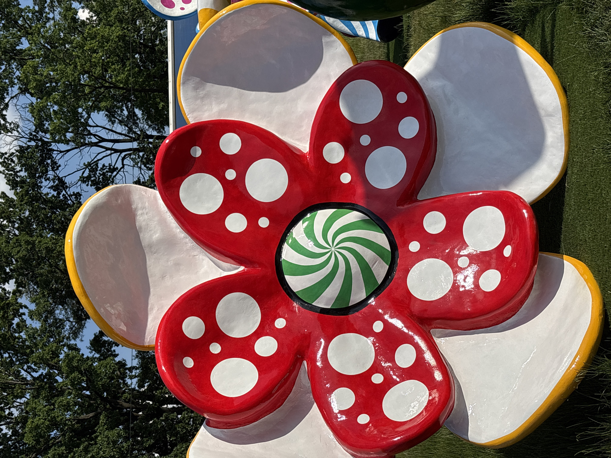 This image showcases a large, vibrant sculpture of a stylized five-petal flower, set outdoors in Hatne, Ukraine. The prominent central flower features glossy red petals adorned with numerous white polka dots of various sizes. Its core is a striking green and white spiral design, reminiscent of a peppermint candy or lollipop. Behind and partially surrounding this main red flower are other petal-like components, primarily white with bright yellow borders, suggesting a larger, whimsical floral art installation.
In the background, the scene reveals lush green trees against a clear blue sky dotted with a few white clouds, indicating a bright, sunny daytime setting. The strong sunlight creates subtle shadows on the sculpture's surfaces, highlighting its three-dimensional form. The overall aesthetic of the sculpture is playful and eye-catching, with a distinct pop-art or cartoonish influence. There are no people visible in the frame, and no discernible text appears on the sculpture or in the background.