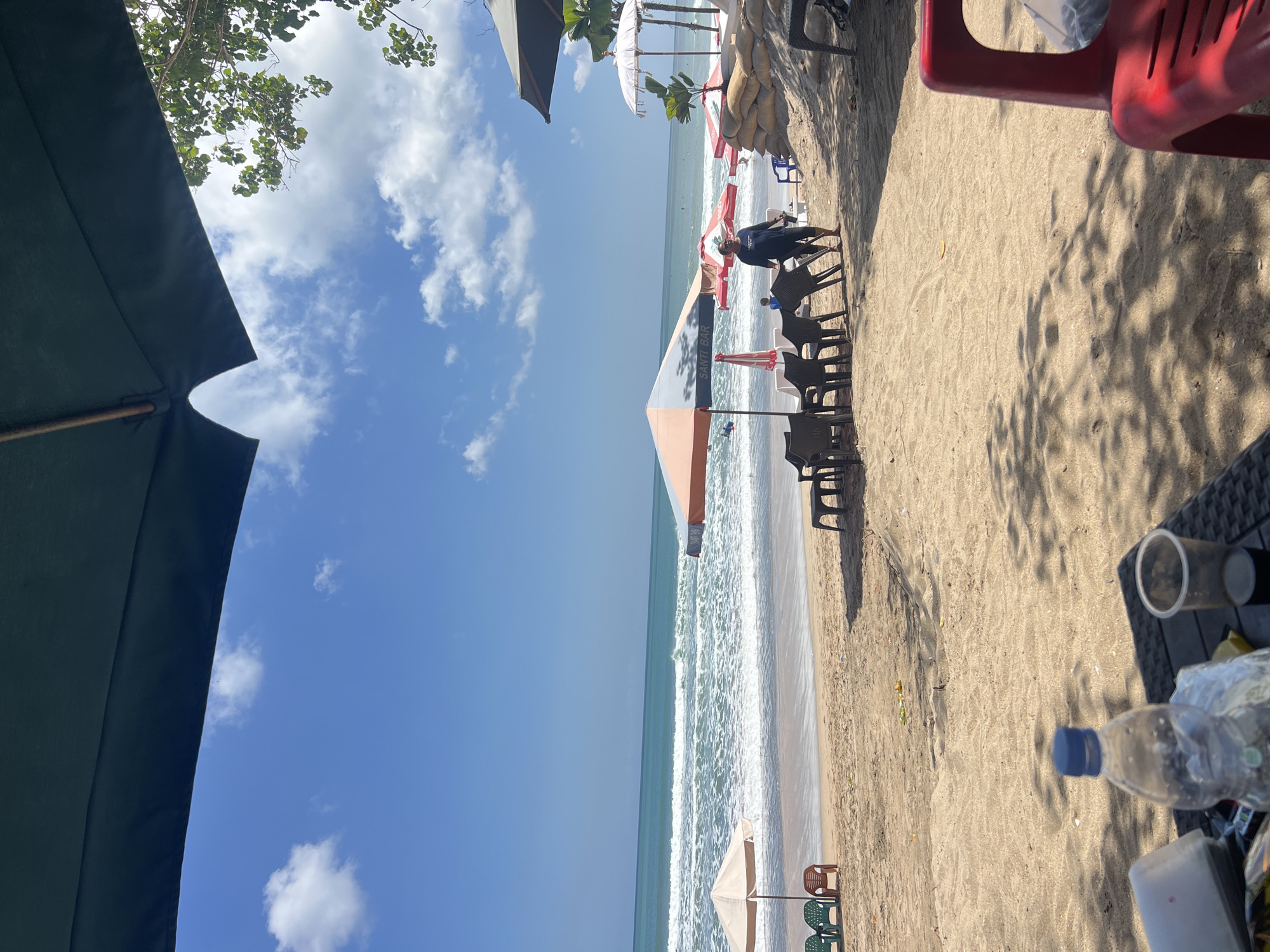 This image depicts a sunny day at Kuta Beach in Bali, Indonesia. The foreground shows a table with a plastic water bottle and a disposable cup, casting dappled shadows on the sand. Beyond the table, the beach is furnished with several sets of chairs and umbrellas. A person in a dark blue t-shirt and dark pants is seen walking among the chairs and umbrellas. Further back, the ocean with gentle waves stretches towards the horizon under a bright blue sky dotted with white clouds. The beach is lined with more umbrellas and what appear to be sandbags, suggesting a defensive structure against the sea. The overall scene conveys a relaxed atmosphere typical of a popular tourist beach.