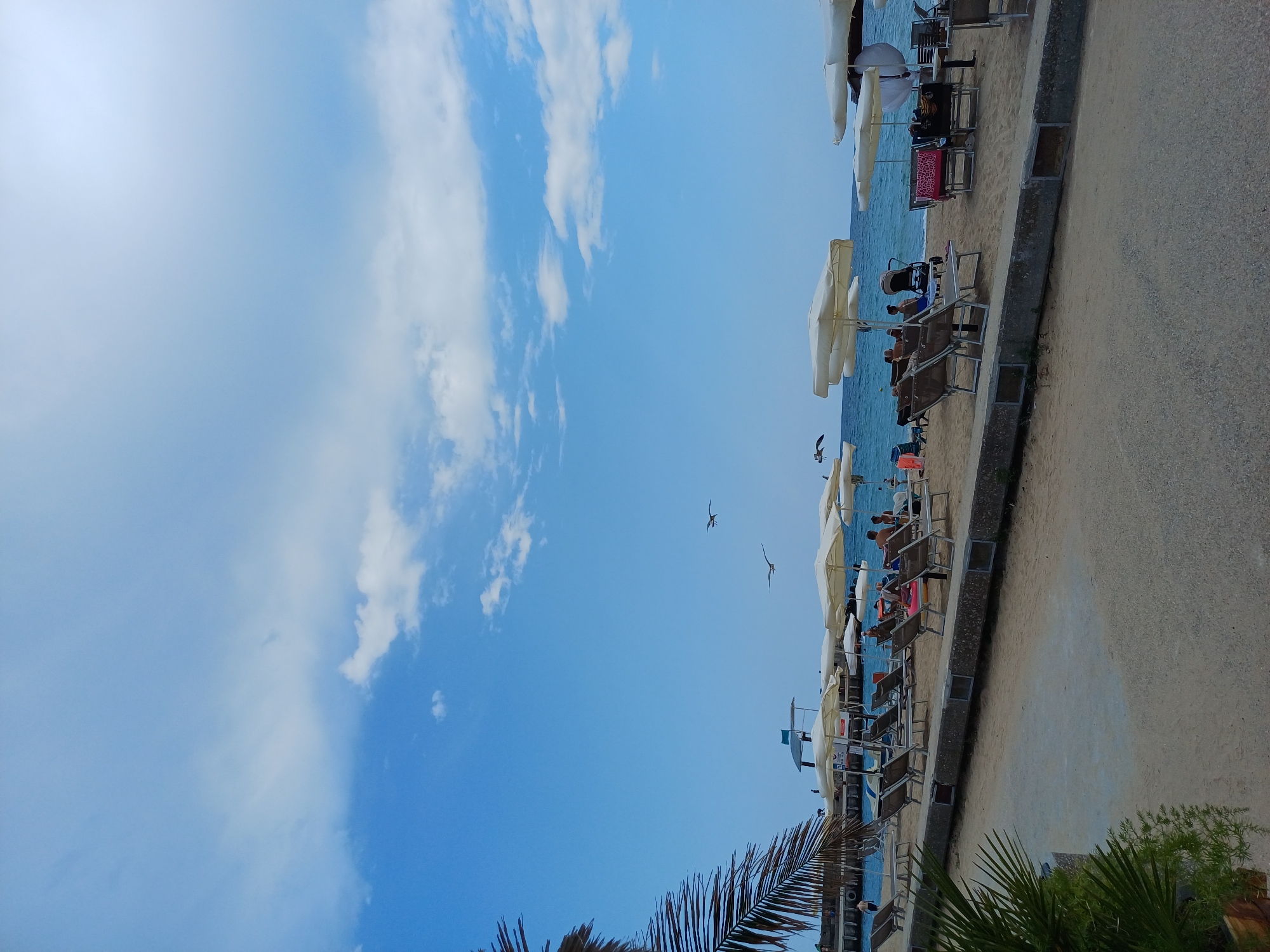 The image depicts a beach scene on a clear day. In the foreground, there is a paved walkway and sand. Further back, there are rows of lounge chairs and umbrellas set up on the sand. People are visible sitting on the chairs and walking around. The sea is visible in the background, with some boats docked. Two seagulls are flying in the sky. A lifeguard tower is also present. The sky is blue with scattered white clouds. Based on the presence of these elements and its typical appearance, this scene is likely located at the Golden Sands beach resort, north of Varna, Bulgaria.