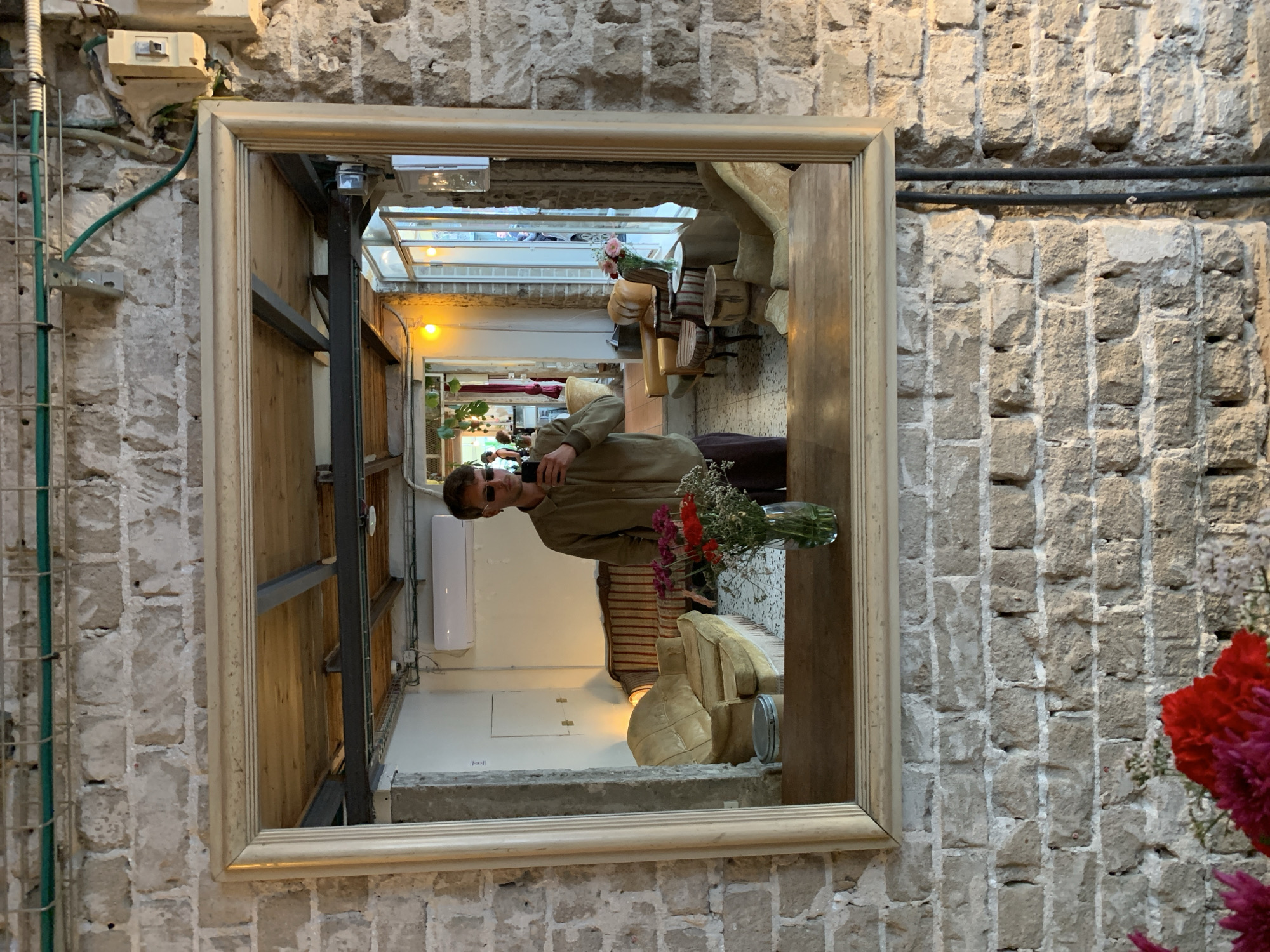 The image shows a rectangular framed mirror hanging on a rough brick wall. Reflected in the mirror is an interior space in what appears to be Tel-Aviv, Israel, indicated by the architecture and design aesthetics typical of the city. The reflection includes:
- A man wearing sunglasses and an olive-green jacket is holding what looks like a mobile phone.
- A vase of flowers sits nearby.
- Seating with cream-colored cushions.
- Architectural details of a building interior with exposed metal framework, suggesting an industrial or loft-style design.
- A skylight structure visible at the top of the frame, giving the interior natural light.
The overall impression is of a modern, design-conscious space, typical of Tel-Aviv's urban aesthetic. The brick wall provides a contrast with the modern elements of the interior.