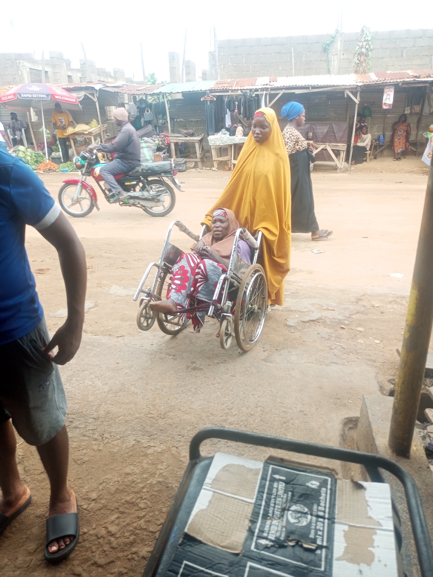 The image, likely taken in Sabon Tasha, Nigeria, shows a woman dressed in a yellow robe pushing an elderly woman in a wheelchair through a bustling outdoor market. The elderly woman is wrapped in patterned fabrics and wears a head covering. The market setting includes stalls with corrugated metal roofs, vendors, and customers. A man on a motorcycle passes by in the background. A person wearing a blue shirt and grey shorts stands in the foreground to the left, with a metal cart carrying a cardboard box visible in the lower right corner. The box is labeled "BENID INDUSTRIES LIMITED" and "ml x 20 x 6 Sachets". The ground appears to be dirt or sand.