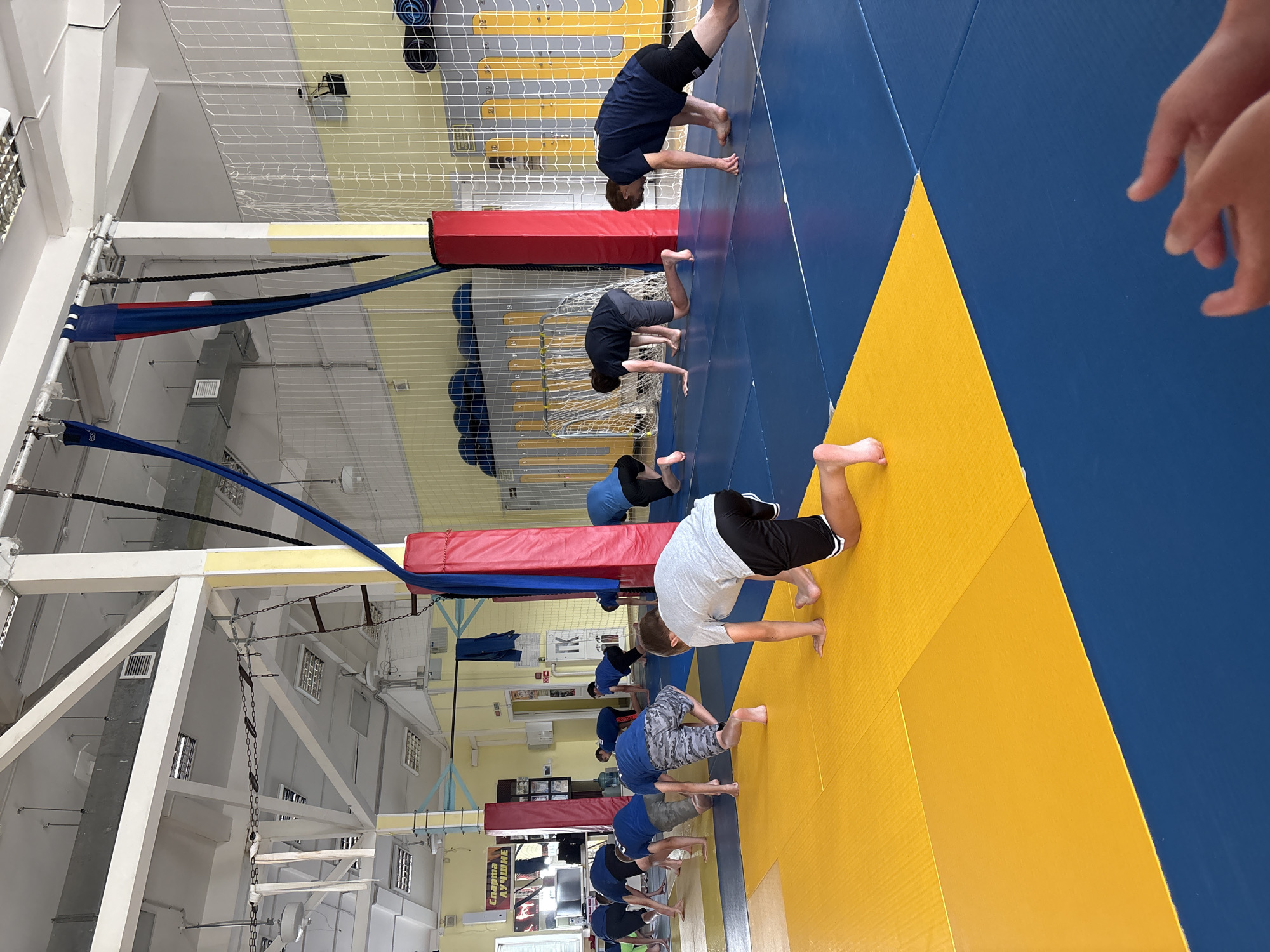 This image shows a group of young males in athletic wear performing an exercise drill on a blue and yellow mat in a gymnasium. The gymnasium appears to be located in Chaikovsky, Russia, indicated by a sign on the wall that reads "СПАРТА ЛУЧШЕ" (Sparta is better) in Cyrillic script. The room has a high ceiling with exposed beams and fluorescent lighting. There are climbing silks hanging from the ceiling, and safety netting lines the walls. Several individuals are in a quadrupedal position, with their hands and knees on the mat, while others are in a similar stance but with their feet further back. The lighting suggests it is daytime.