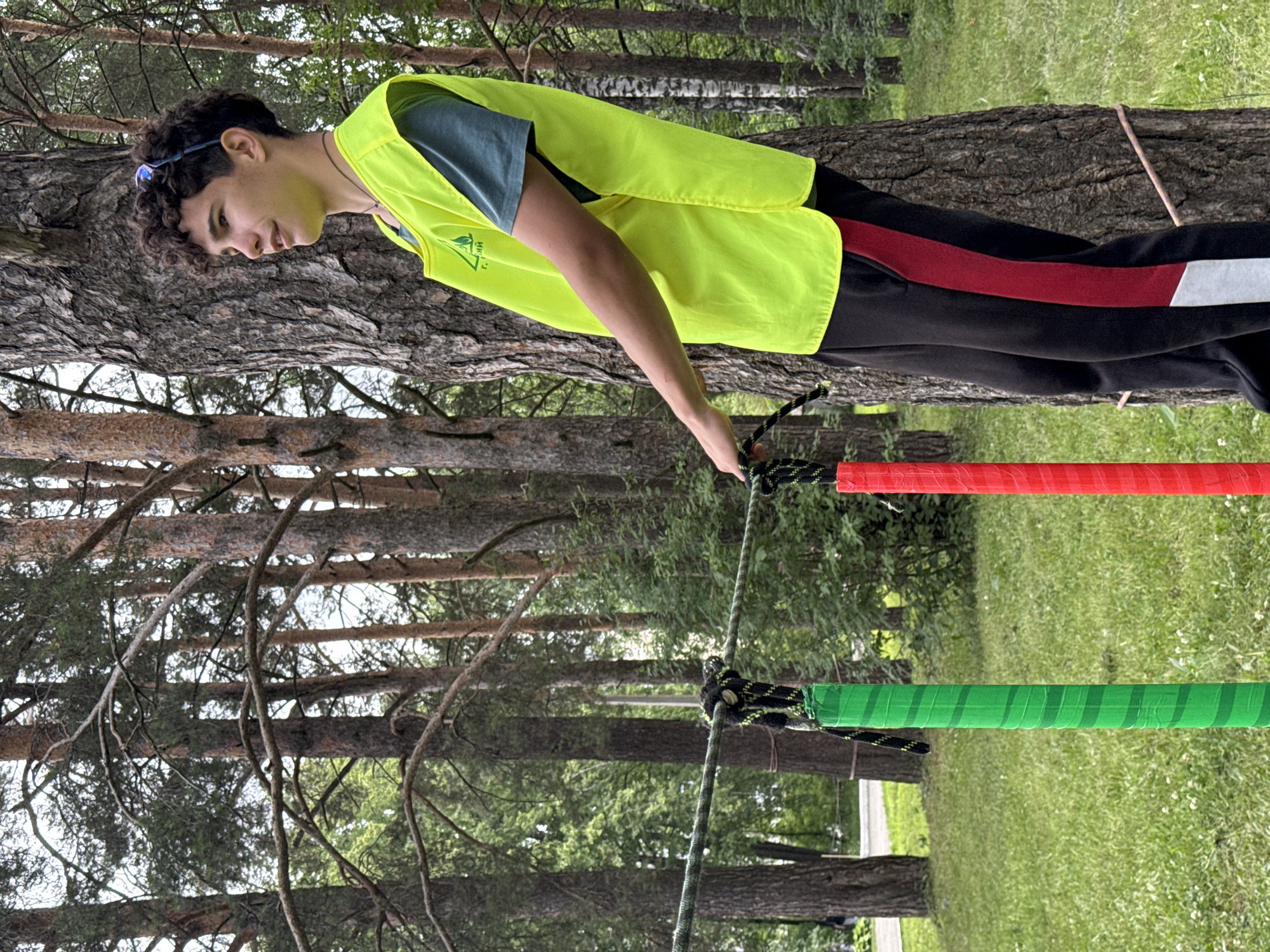 A young male with curly dark hair and sunglasses perched on his head stands next to a large tree, wearing a bright yellow safety vest over a dark blue t-shirt and black track pants with a red stripe. He is holding onto a rope that stretches horizontally across the frame, secured by a knot. In the foreground, a green pole with dark stripes and a red pole with dark stripes are visible, also connected to the rope. The background is a forest scene with tall trees and green grass, suggesting an outdoor recreational area. The image appears to have been taken in the city of Chaikovsky, Russia, likely at an adventure park or ropes course.