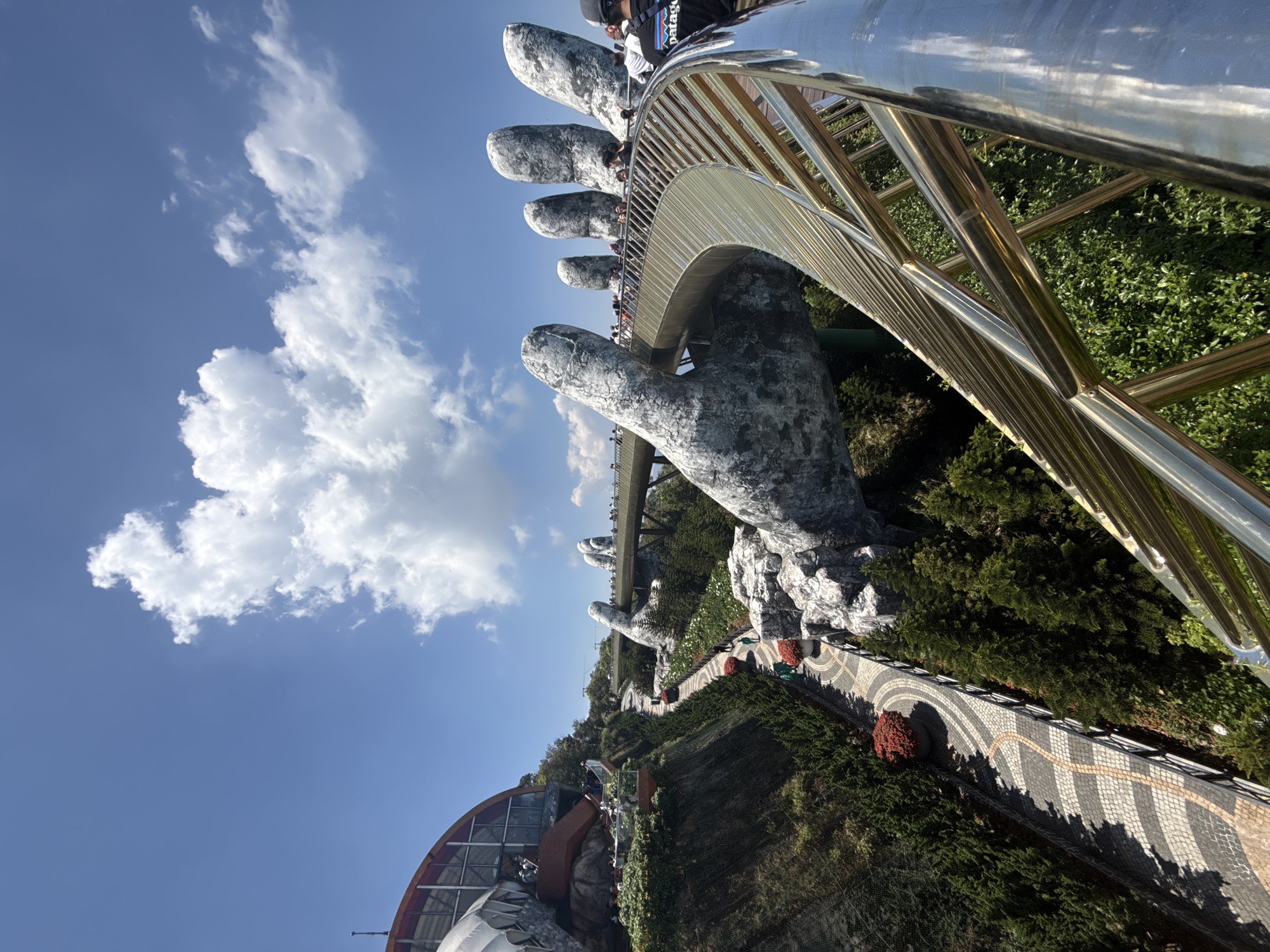 The image captures the Golden Bridge in the Bà Nà Hills near Đà Nẵng, Vietnam. The bridge is supported by two large, sculpted stone hands. People are walking along the bridge. The bridge has gold-colored railings. Lush greenery surrounds the structure, and a blue sky with white clouds is in the background. Part of a building is visible in the lower left corner. A patterned pathway borders the green landscape.