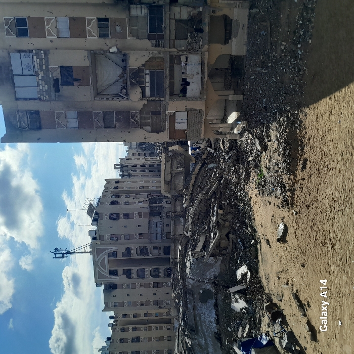 The image depicts a scene of extensive urban destruction in Khan Yunis, Palestinian Territory, under a partly cloudy sky. In the foreground, a field of disturbed earth, sand, and various sizes of rubble and debris stretches across the lower portion of the frame. A blue tarp or fabric is visible on the far left amidst the debris.
The midground features a substantial mound of collapsed building materials, including broken concrete slabs, twisted rebar, and shattered masonry, indicating the complete destruction of a structure.
In the background, multiple multi-story buildings are visible. To the immediate right, a heavily damaged residential building stands, with numerous sections of its facade and walls missing, revealing interiors. Windows are shattered or entirely absent, with some openings covered by makeshift materials like white fabric or cardboard. The concrete exterior walls display widespread pockmarks and holes consistent with projectile impacts. On one of the middle floors, clothes are visible hanging within a damaged apartment. The ground floor exhibits arched openings.
Further in the center and to the left, a series of similar multi-story concrete buildings appear damaged, though less severely than the structure on the right. These buildings feature balconies and architectural details, with many windows broken or missing. A prominent central building has a cellular communication tower mounted on its roof, alongside what appear to be solar panels. No people are visible within the scene, and the overall environment is dry and dusty. The bottom-left corner of the image contains a "Galaxy A14" watermark.
