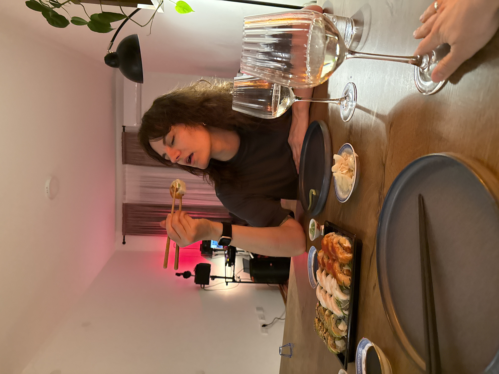A woman with dark, curly hair, wearing a grey t-shirt and a wristwatch, is eating sushi with chopsticks at a table. Two wine glasses filled with white wine are on the table, along with sushi platters, small dishes of ginger, and plates. The scene appears to be indoors, with a black lamp overhead and sound equipment visible in the background. A window with closed shades is present. The location is unknown.