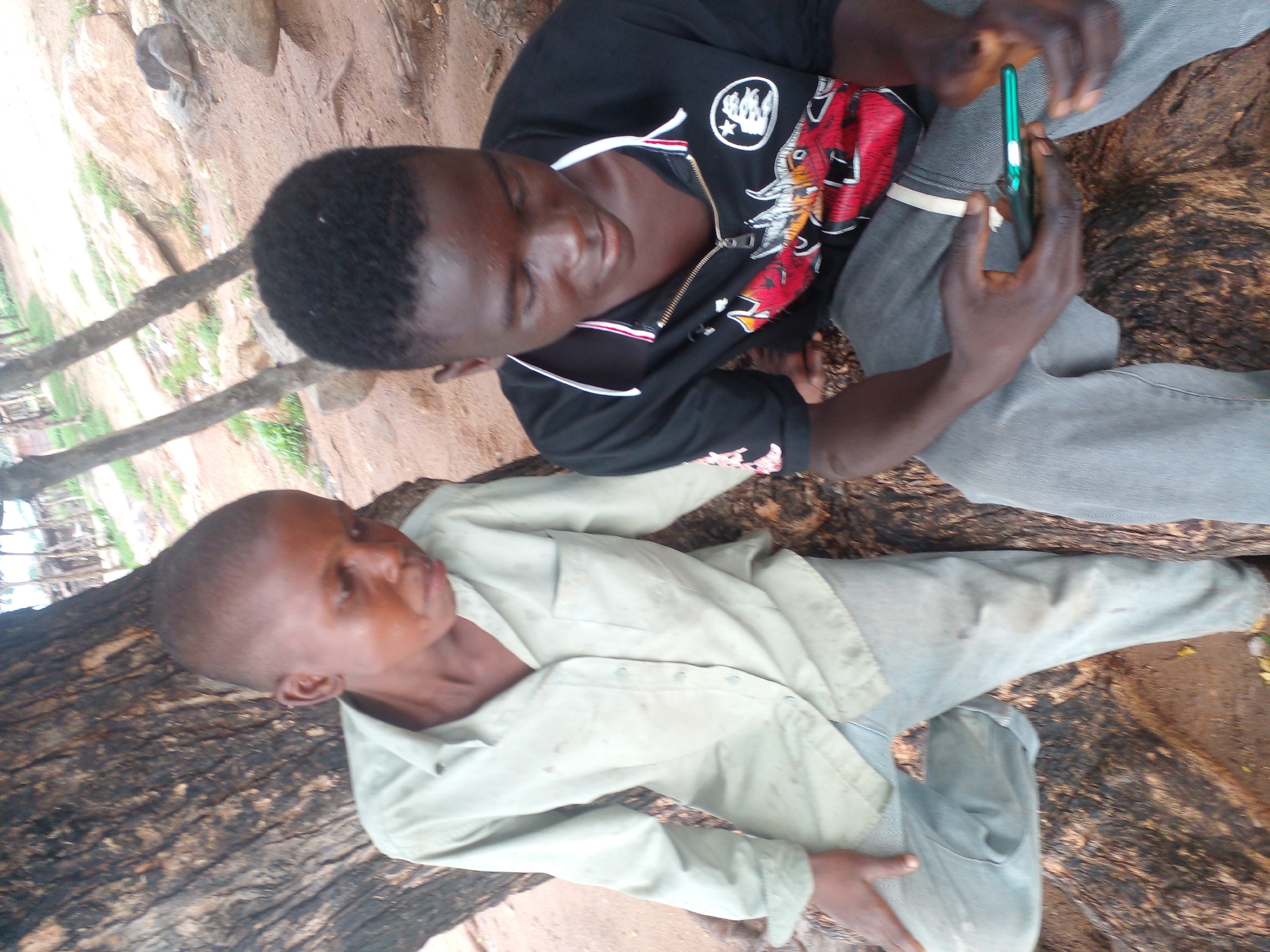 Two young males are seated outdoors, leaning against the trunk of a large tree. The male on the left wears a light-colored, long-sleeved button-down shirt and light-colored pants. He has a short haircut and is looking down and to the right. The male on the right wears a black t-shirt with a graphic design on the front, featuring a red, white, and black dragon-like image and a white circular emblem. He also wears grey pants and is looking down at a green smartphone he is holding in his hands. The environment appears to be rural or semi-rural, with sandy ground, rocks, and sparse vegetation visible in the background. The image was taken in Langtang, Nigeria.