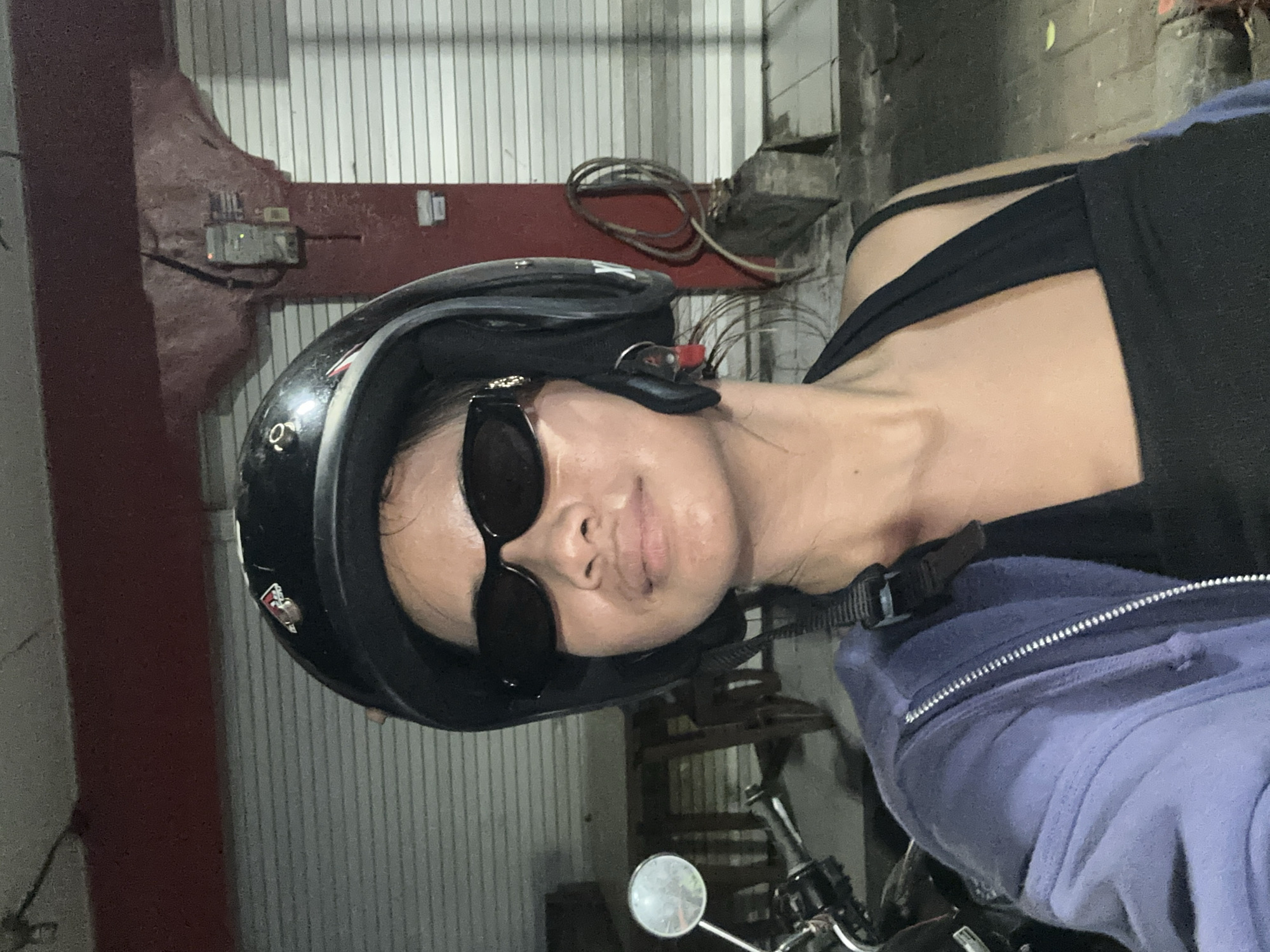 The image depicts a young woman wearing a black helmet and sunglasses, seated on a motorcycle, and taking a selfie in what appears to be an urban environment in Denpasar, Indonesia. She is wearing a dark tank top and a light purple zip-up hoodie. The background shows a weathered red wall, a closed corrugated metal shutter, and a rolled-up hose. The lighting suggests it is either dusk or nighttime.