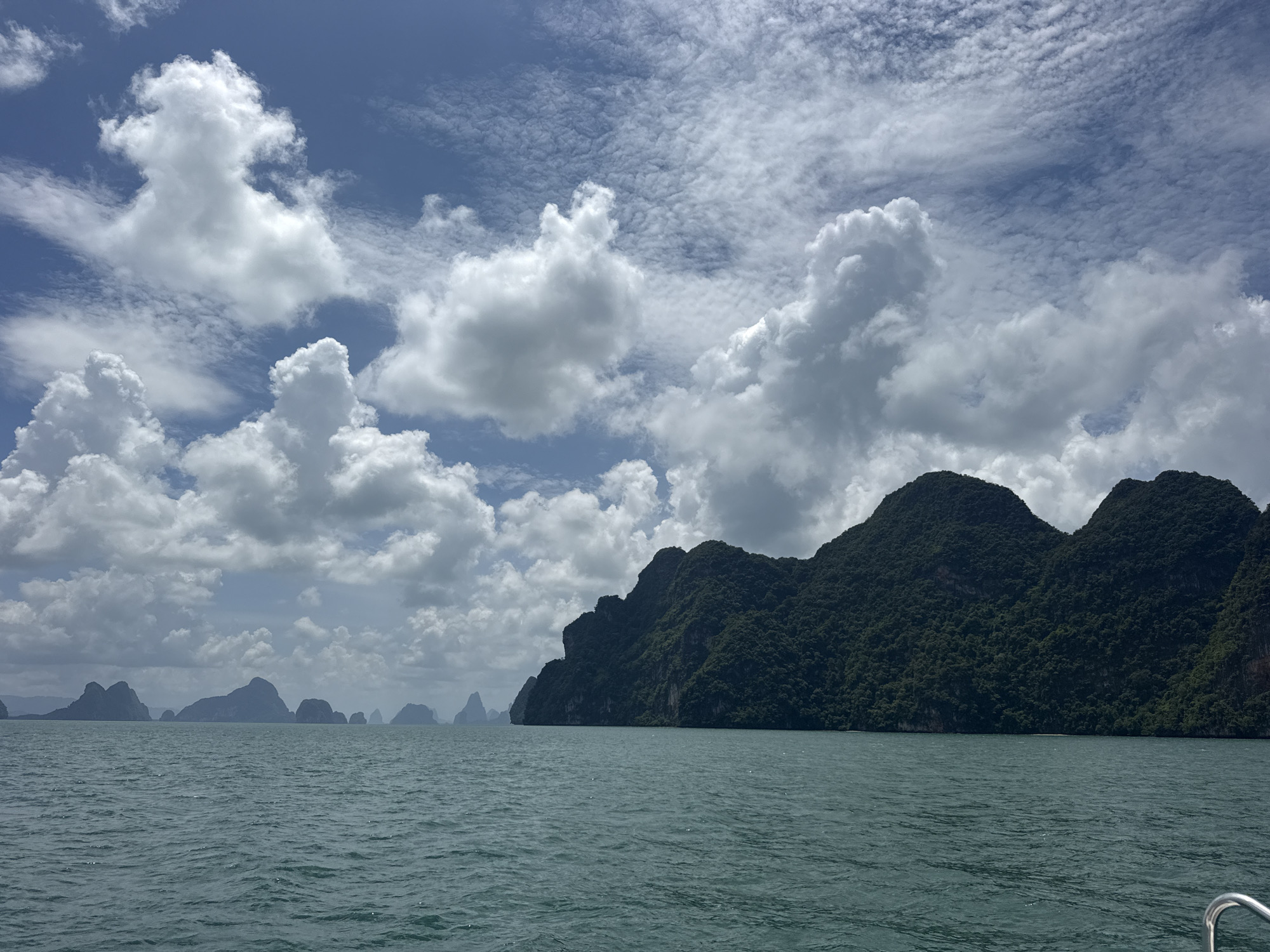 The image depicts a serene seascape with lush, mountainous islands under a partly cloudy sky. The water is a calm, teal-green, reflecting the sky and clouds. In the foreground, the choppy surface of the ocean suggests movement, possibly from a boat. The middle ground is dominated by several large, densely forested islands with steep, rocky slopes. In the distance, a chain of smaller islands stretches across the horizon. The sky is a vibrant blue, populated with large, fluffy white cumulus clouds and smaller, wispy cirrus clouds. The scene is a picturesque representation of the natural beauty of the region, likely captured from a moving vessel. This specific landscape is characteristic of Phang Nga Bay, near Krabi and Phuket, in Thailand, and while Krasom is a locality, it is not a city and this image does not depict it.