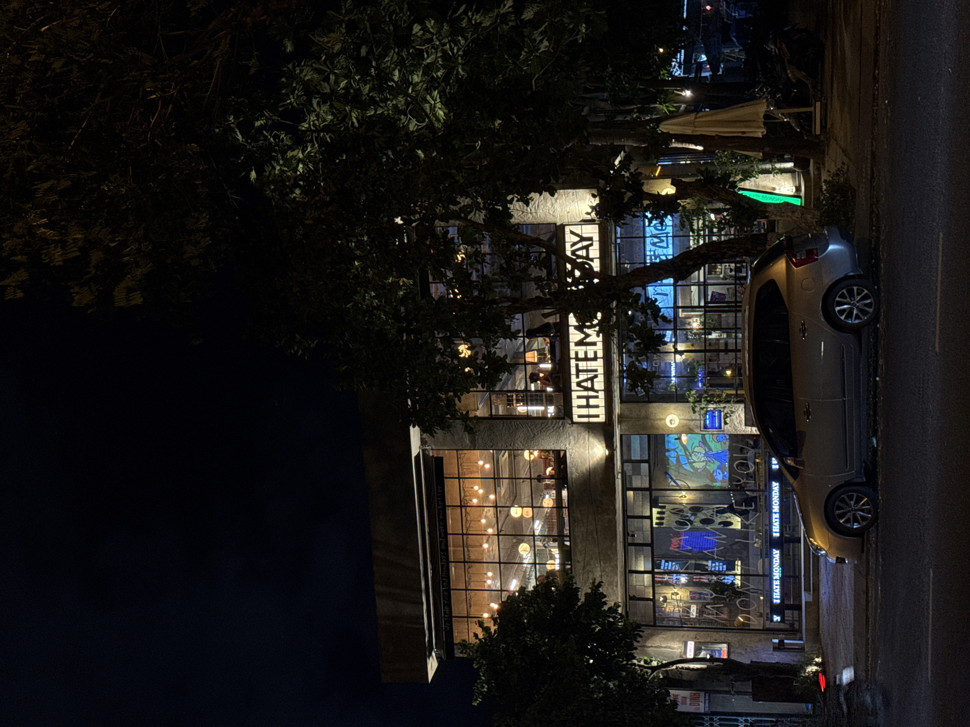 The image shows a building, possibly a cafe or restaurant, at night. A gray car is parked on the street beside the building. The building has a multi-story facade with large windows that reveal an illuminated interior with people inside. Several illuminated signs can be seen on the building, including "I HATE MONDAY," and "THATEM DAY". Trees partially obscure the building. Based on OCR of the image, the building is located in the city of Đà Nẵng, Vietnam.
