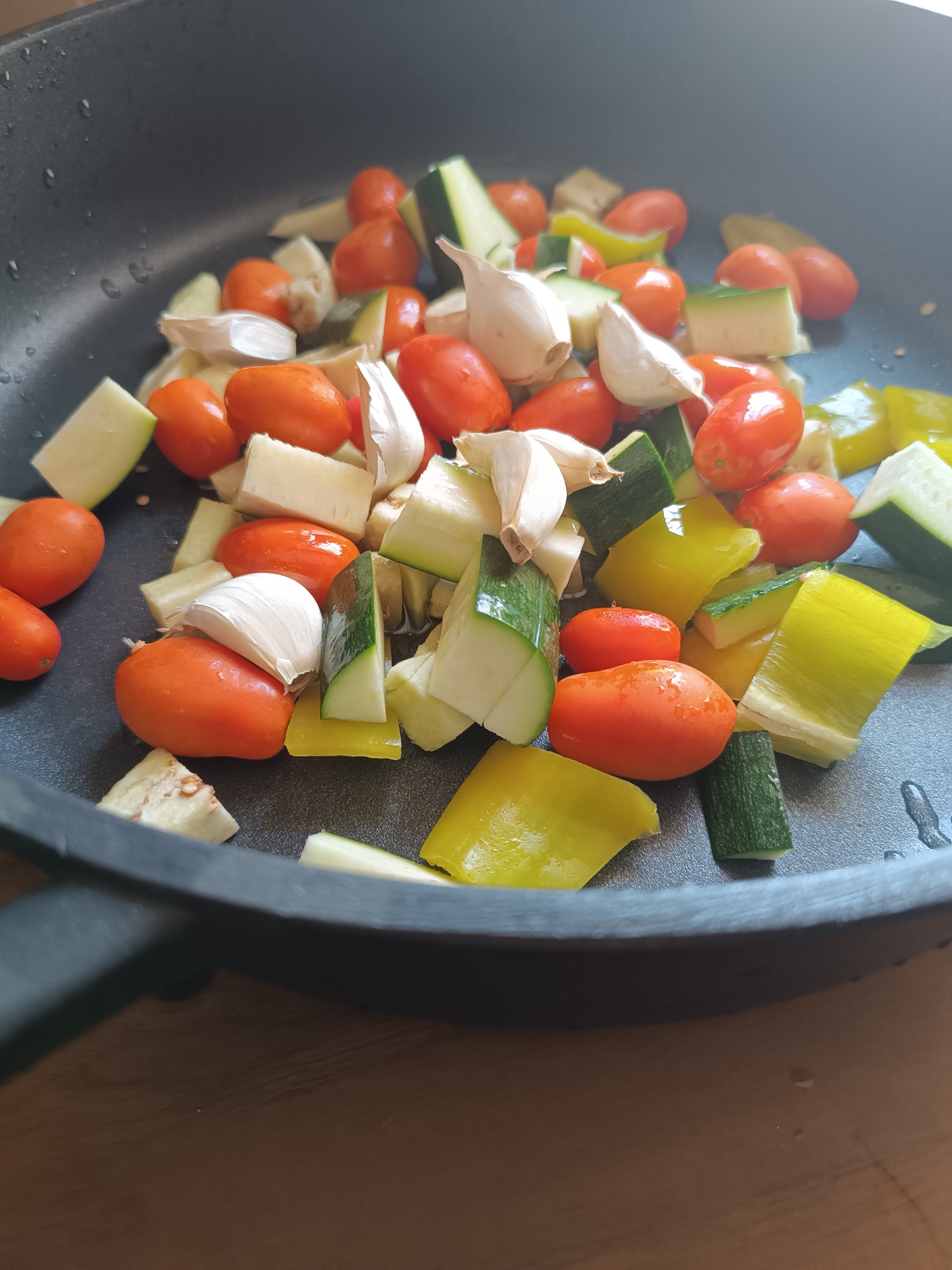 A close-up, high-angle shot shows a variety of chopped vegetables and garlic cloves in a black non-stick frying pan. The vegetables include small, whole, red cherry tomatoes, quartered zucchini, chopped yellow bell pepper, and sliced leeks. Several cloves of unpeeled garlic are also visible. The vegetables are arranged somewhat haphazardly in the pan, which is placed on a dark wooden surface. The scene is lit from the left, casting shadows and highlighting the textures of the ingredients. The presence of water droplets on the pan suggests that the vegetables may have been recently washed or are about to be cooked. There is no indication of any specific location within the city of Viadana, Italy, or any human activity.