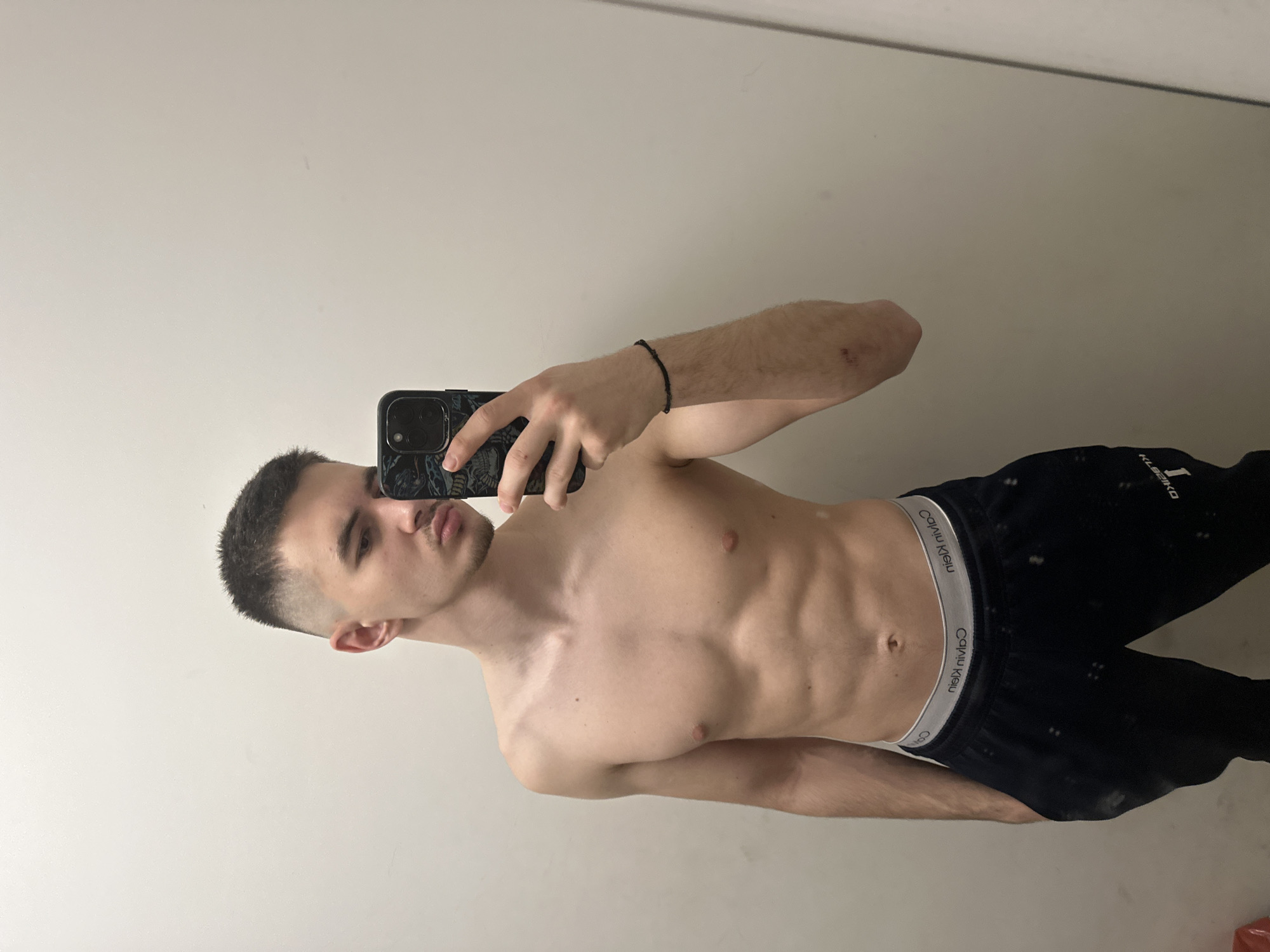 A young man is taking a selfie in what appears to be an indoor location with a plain, white wall. He has short, dark hair and is shirtless, wearing black sweatpants with a Calvin Klein waistband. He's holding a black smartphone in his right hand, covering part of his face. The lighting is neutral, and the image is oriented vertically. The location is not discernible as Sofia, Bulgaria, based on the visual information provided.