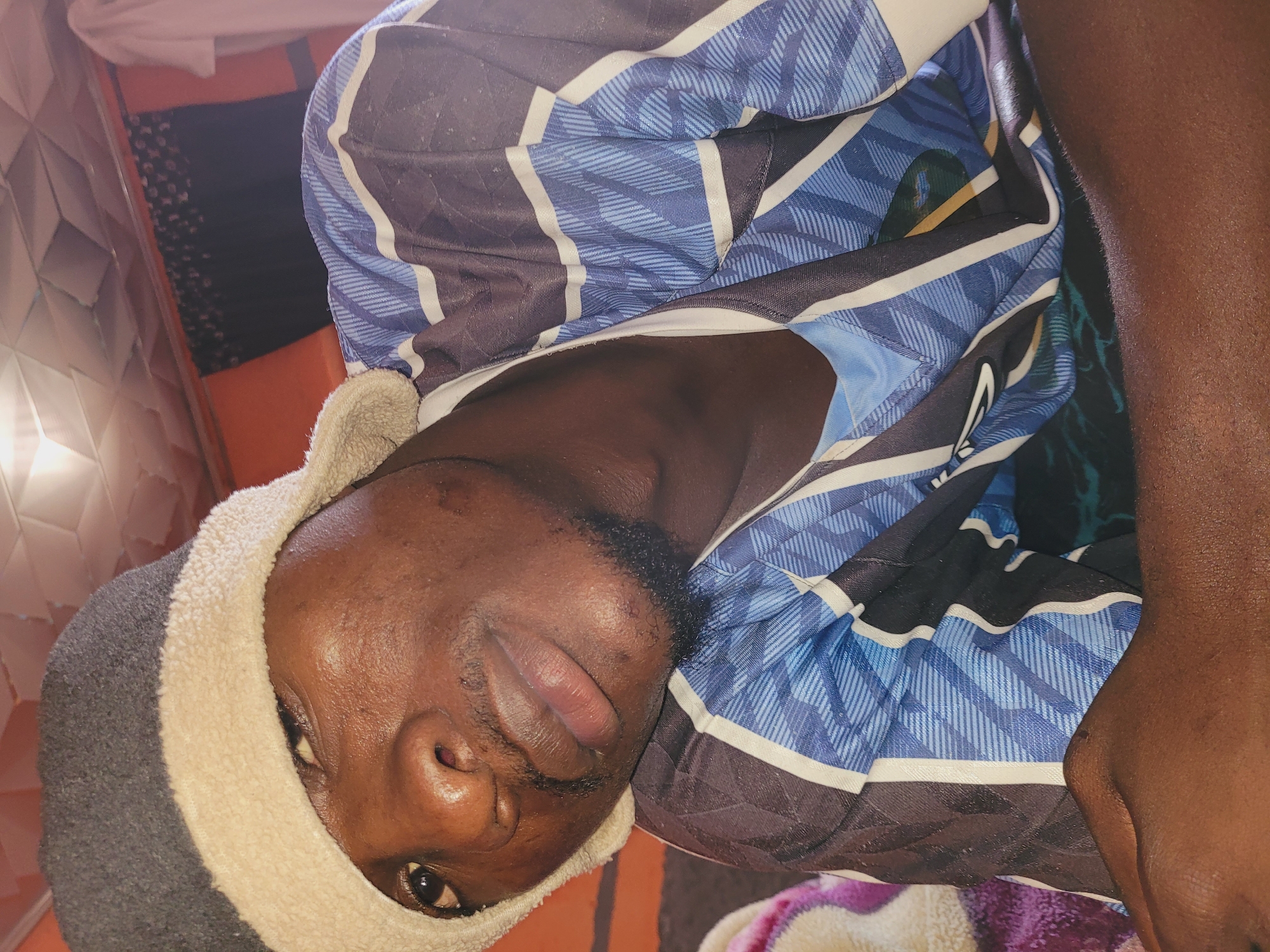 In Bukuru, Nigeria, a close-up, indoor, eye-level shot captures a young Black male resting on a bed, wearing a gray and cream beanie and a blue, white, and black striped t-shirt. His head is tilted, and he is looking towards the camera with a relaxed expression. The background features a ceiling with diamond-shaped panels, orange and black curtains, and a white garment hanging. The scene is illuminated by overhead lighting, creating shadows and highlights on the subject's face and the surrounding environment.