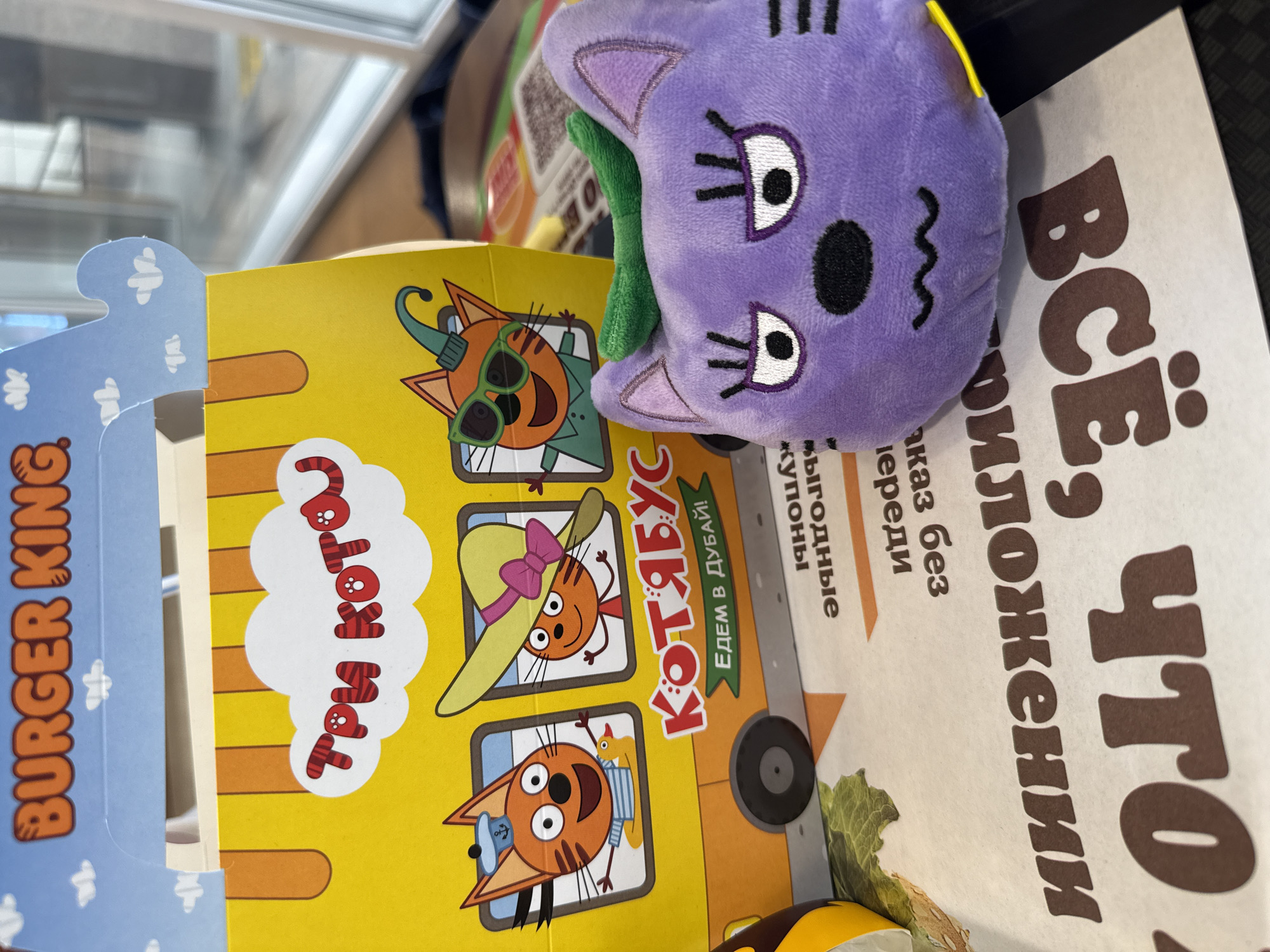 An indoor scene, likely a fast-food restaurant in Tobolsk, Russia, features a purple plush toy and various cardboard promotional materials. The main subject is a purple, plush cat-like toy with large white eyes, black pupils, embroidered eyelashes, a black oval nose, and a wavy black embroidered mouth. It has pointed ears and a small green stem-like element on top, resembling an eggplant or aubergine.
The toy rests on a yellow cardboard box that features illustrations of orange cats from the Russian animated series "Три Кота" (Three Cats). The box is designed as a "КОТЯБУС" (Kotyabus, a portmanteau of "cat" and "bus") with a visible black wheel. Illustrations on the box show three distinct cat characters: one wearing a green conical hat and sunglasses, another in a yellow wide-brimmed hat with a pink bow, and a third in a blue sailor hat. Text on the box includes "Три Кота", "КОТЯБУС", and "ЕДЕМ В ДУБАЙ!" (We're going to Dubai!).
Adjacent to the yellow box is a blue cardboard piece displaying the "BURGER KING" logo, indicating the restaurant's brand. Also visible is a white sheet of paper or cardboard with large brown Russian text. This text reads "ВСЁ, ЧТО" (All that) and includes phrases like "Филожении" (likely part of "Приложении" - in the application), "заказ без очереди" (order without queue), and "ВЫГОДНые купоны" (profitable coupons), suggesting a promotion for Burger King's mobile app or deals. The background shows reflections of what appears to be a glass surface or window. No specific activity is depicted, but the items suggest a purchase or meal at Burger King, possibly a kids' meal with a toy.