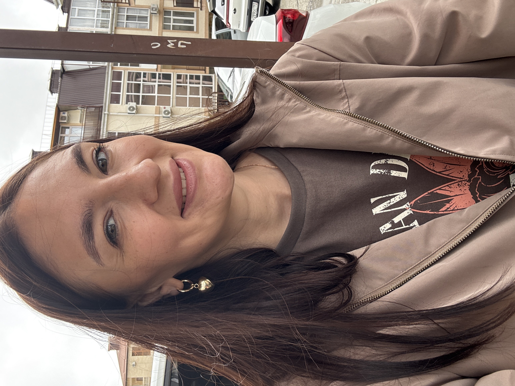 A young woman is seen in a close-up shot, smiling slightly, facing the camera. She has dark hair, fair skin, and is wearing a brown t-shirt and a beige jacket. She is also wearing gold earrings. The background consists of buildings and cars. The image was taken in Sochi, Russia, as indicated by the Cyrillic writing on a sign in the background.