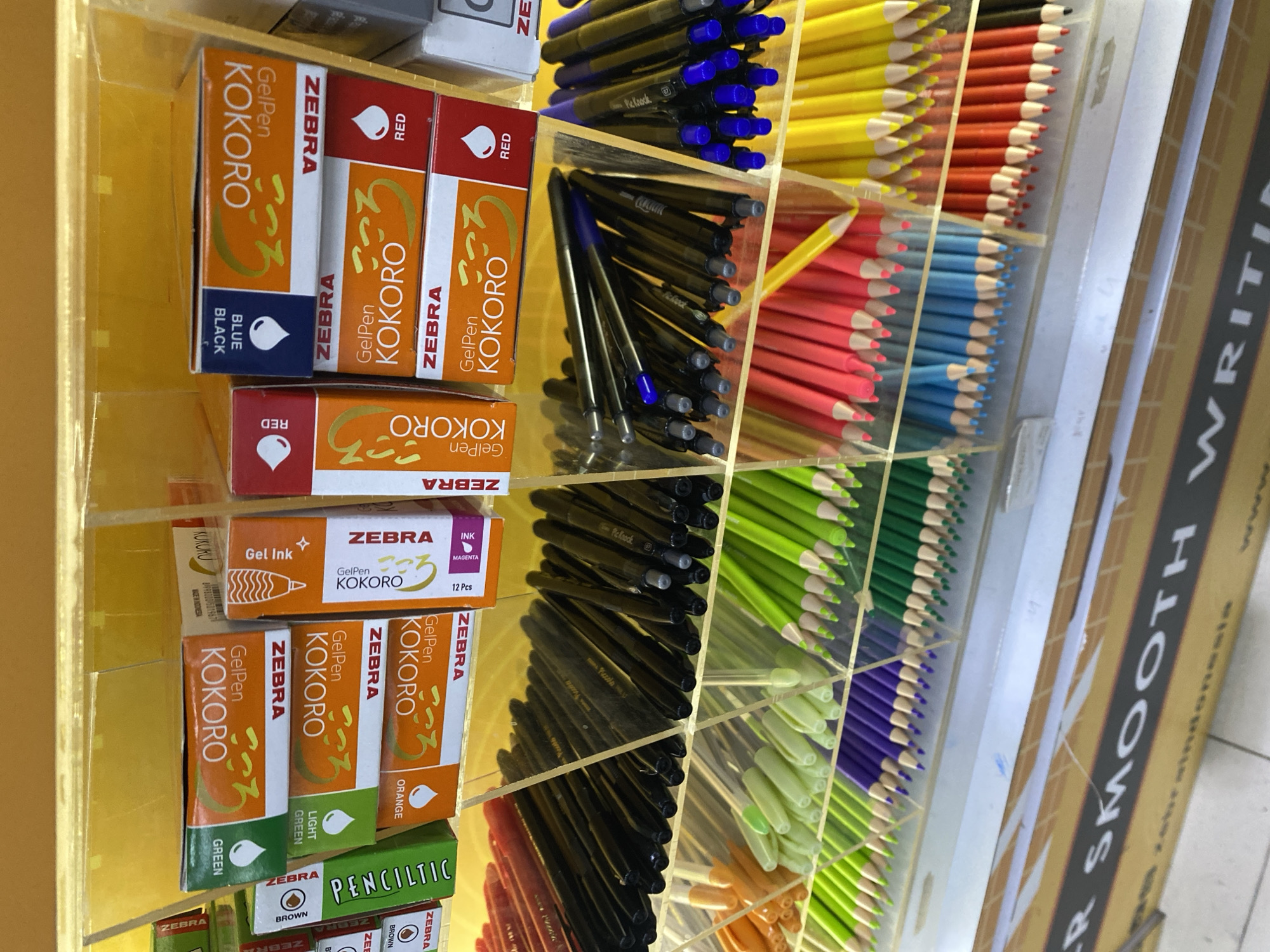 The image displays a retail shelf filled with various writing and drawing supplies, likely in a stationery store. Prominently featured are boxes of Zebra brand gel pens, specifically the "Kokoro" line, in different colors like green, orange, blue, and red. Below the pens, shelves are stocked with colored pencils in vibrant hues such as neon green, purple, pink, yellow, orange, and red. The background shows a yellow display wall. The bottom portion of the image reveals a cardboard display box with text that includes "SMOOTH WRITING" and "Zebraindonesia," suggesting the location is a Zebra authorized retailer in Depok, Indonesia. No people are visible in the frame, and the lighting suggests an indoor retail environment.