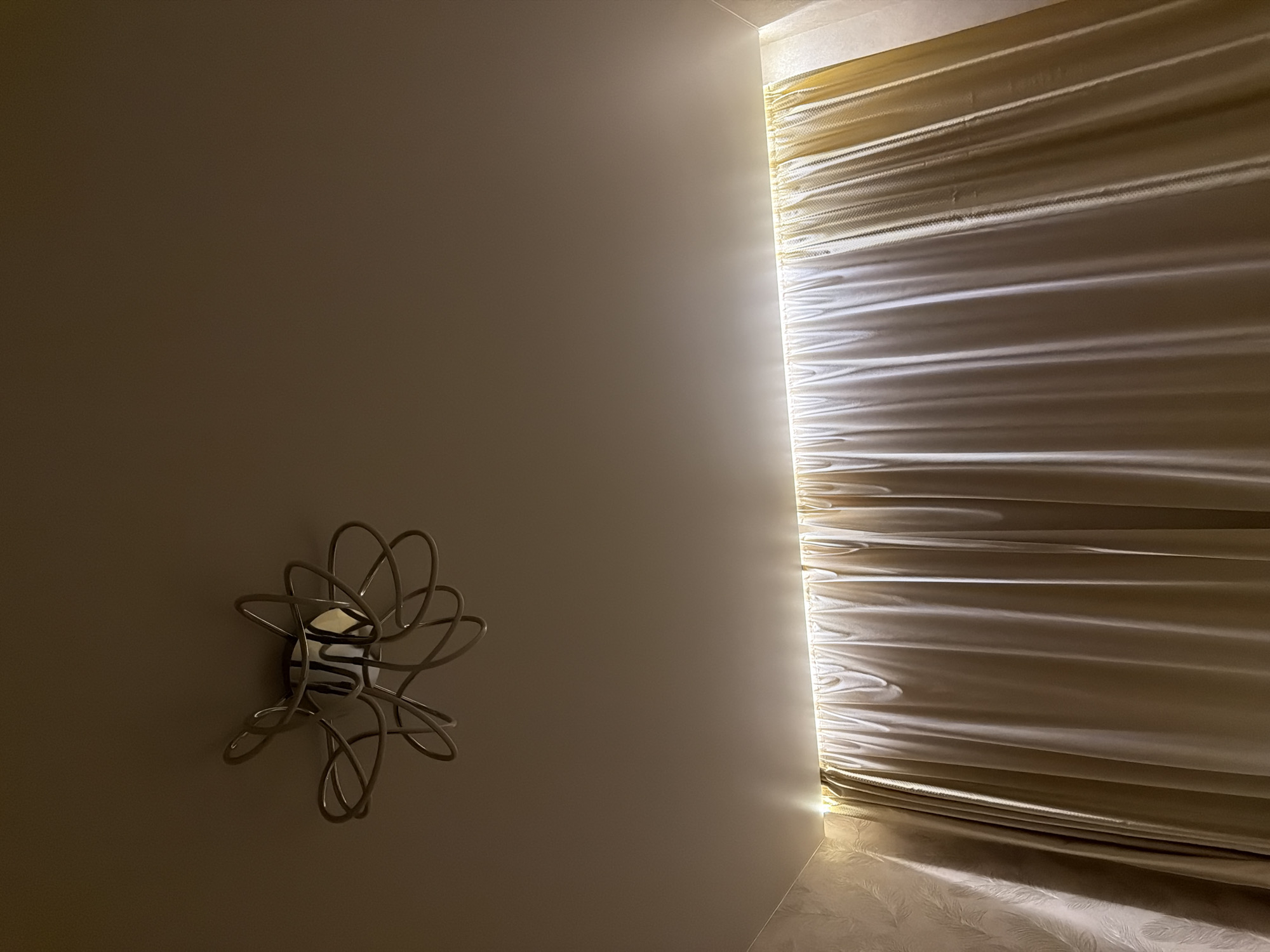The image displays an indoor scene, likely a room in Izhevsk, Russia, characterized by warm, subdued lighting. On the right side, a window is covered by cream or off-white blinds or shades, possibly Roman blinds, which are strongly backlit by a hidden light source. This backlighting creates distinct horizontal bands of light and shadow across the fabric of the blinds, highlighting their texture and folds. A bright, warm glow emanates from behind the blinds, illuminating the adjacent wall and the textured surface directly beneath the window, which features a subtle, possibly feathered or leafy pattern.
The left side of the frame is dominated by a smooth, light brown or greige wall. Mounted on this wall, slightly to the left of the center, is a decorative metallic sconce or fixture. It features an intricate, abstract design composed of intertwining loops and swirls, resembling a stylized flower or starburst. A reflective surface or a small light bulb can be seen at its center, catching some of the room's ambient light.
The overall atmosphere is calm and ambient, suggesting either evening or night with artificial lighting, or perhaps heavily filtered daylight. No people or visible text are present in the image.