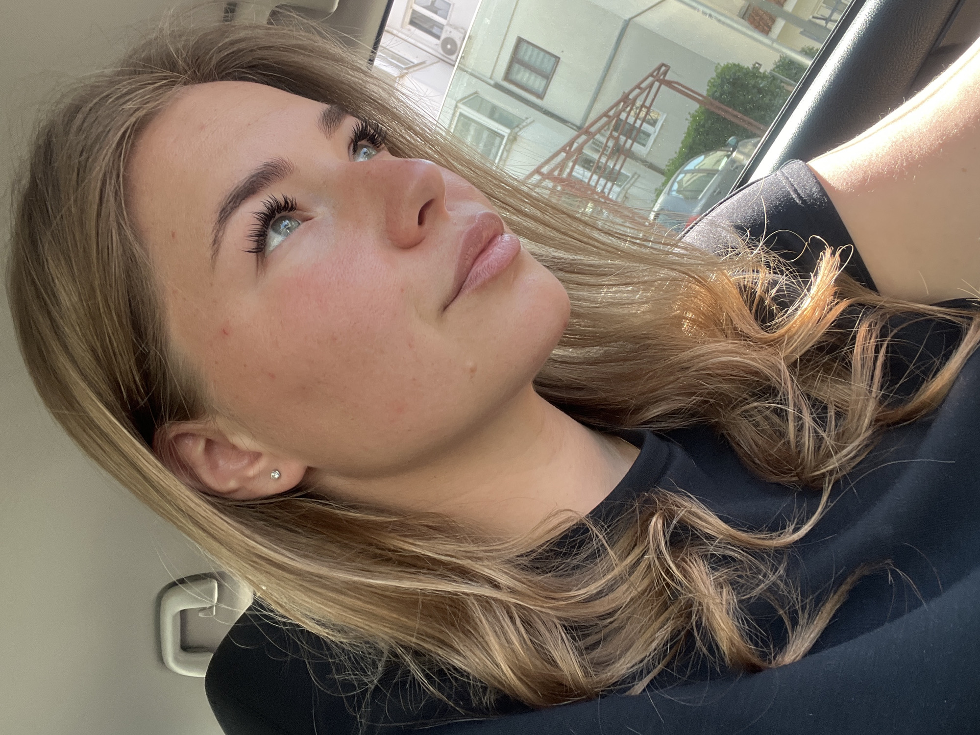The image depicts a female individual, approximately from the chest up, seated inside a vehicle during daylight hours in Budva, Montenegro. The individual has light skin, blue eyes, and light brown to blonde wavy hair, parted on the right. She wears a dark, likely black, short-sleeved garment. A small, silver-colored stud earring is visible in her right earlobe. Her gaze is directed towards the right, out of the vehicle's window. Minor facial blemishes are visible on her forehead and cheek. The vehicle's interior includes a light-colored headliner and a white grab handle positioned above and to the left of the individual. Outside the window, a multi-story, light-colored building with multiple windows is visible, along with an external air conditioning unit. A dark-colored vehicle, green foliage, and a reddish-brown metal structure are also present in the background. Sunlight illuminates the individual's face and hair from the right.