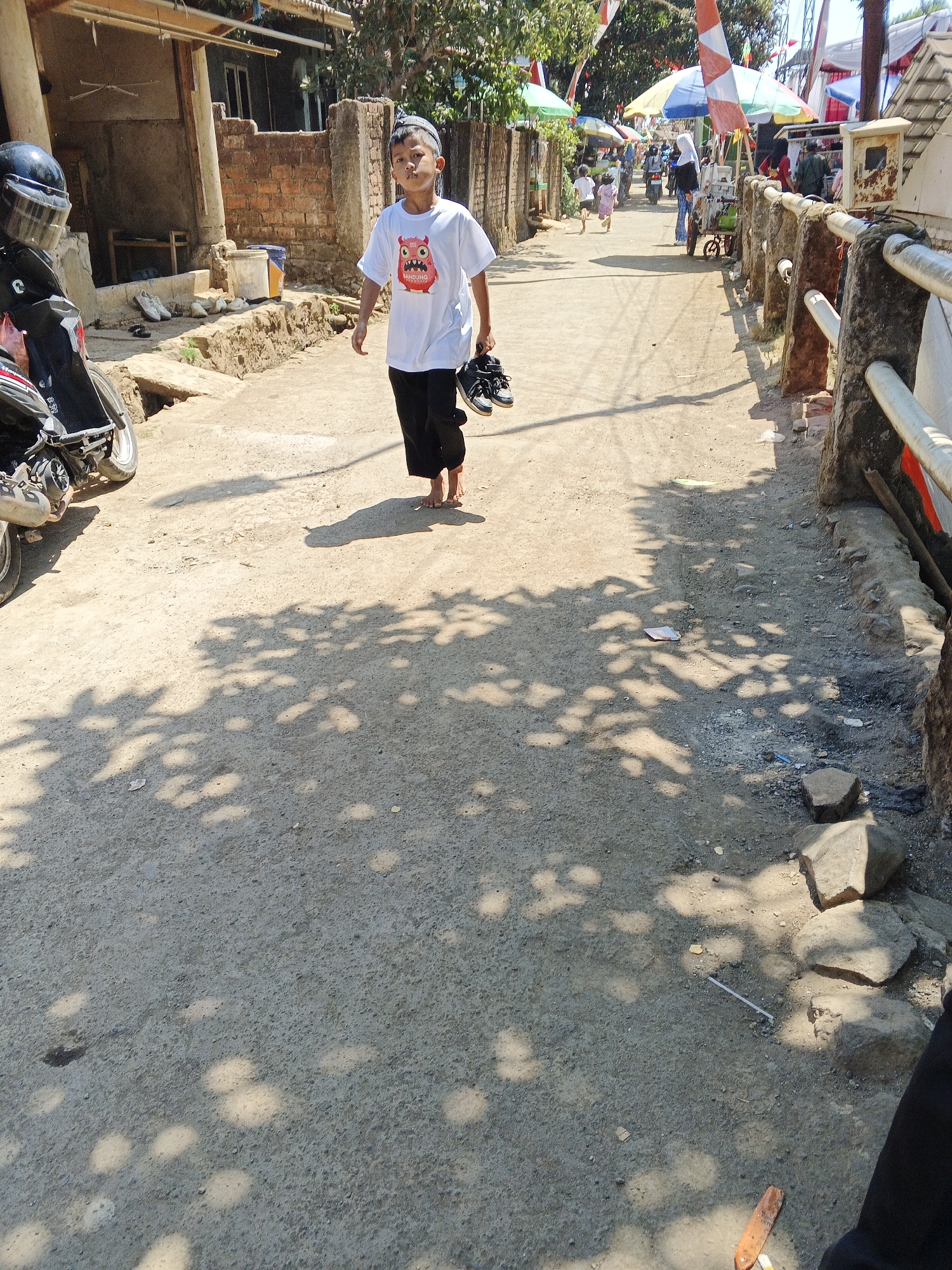 The image depicts a street scene in Sukaharja, Indonesia, on a sunny day. A young boy, wearing a white t-shirt with a cartoon owl graphic and black pants, is walking on a dirt and gravel path. He is barefoot and carrying a pair of black sneakers in his right hand. To his left, a dark-colored motorcycle is parked. The background shows a rustic brick building and several market stalls with colorful umbrellas. Other people are visible in the distance, including children running and adults walking. Shadows from trees and structures are cast across the path. The environment appears to be a typical village or small town street with elements of local commerce.