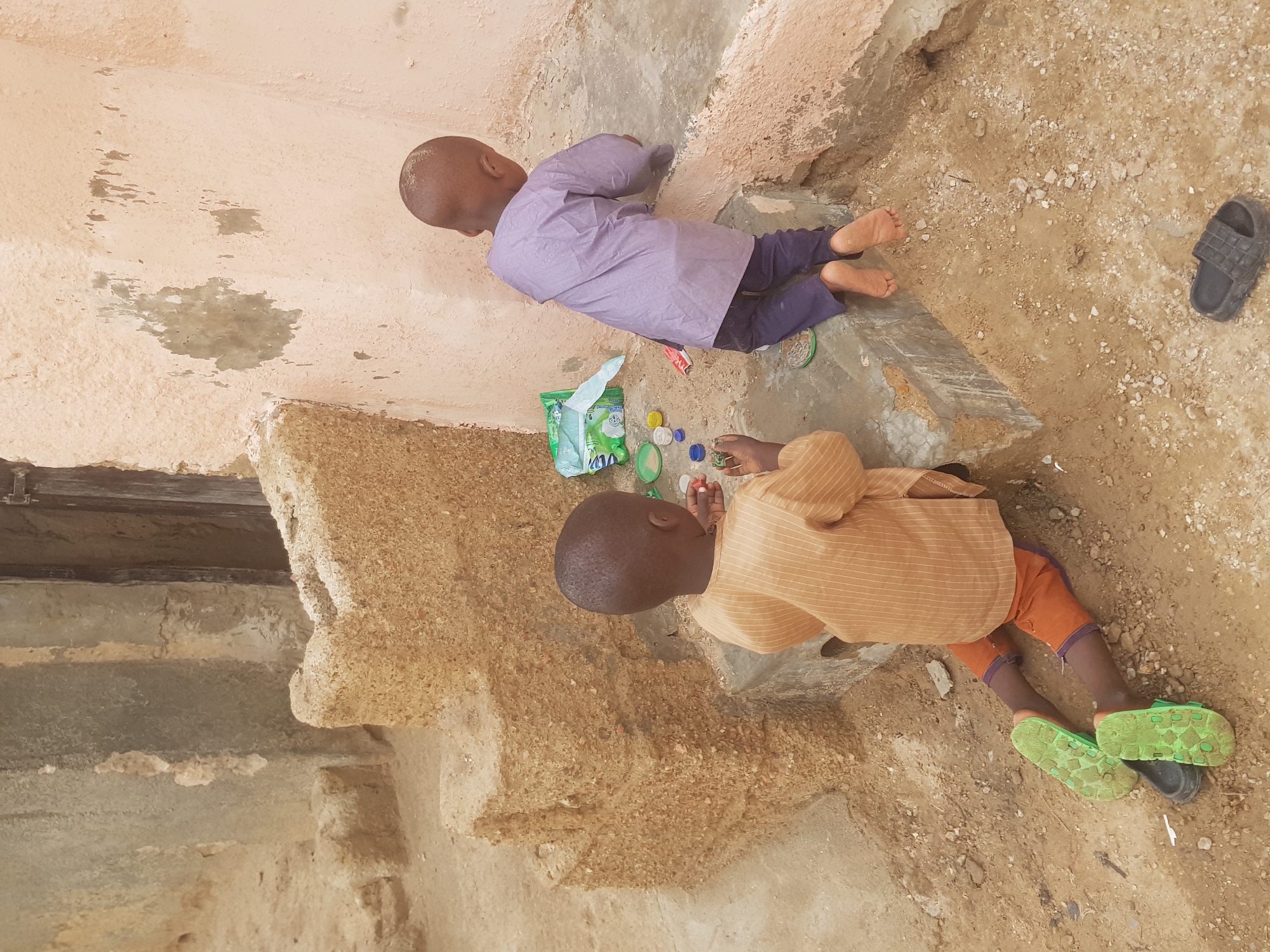 Two young boys are lying on the ground, playing with bottle caps in what appears to be an outdoor area in Dutsinma B, Nigeria. One boy wears a light purple shirt and dark pants, lying barefoot. The other boy wears a tan, striped shirt and orange shorts, with mismatched green and black sandals. Nearby are a package of wipes and several scattered bottle caps. A single black sandal lies near the first boy. The area is a mix of concrete and dirt, with a wall and other architectural elements visible in the background.