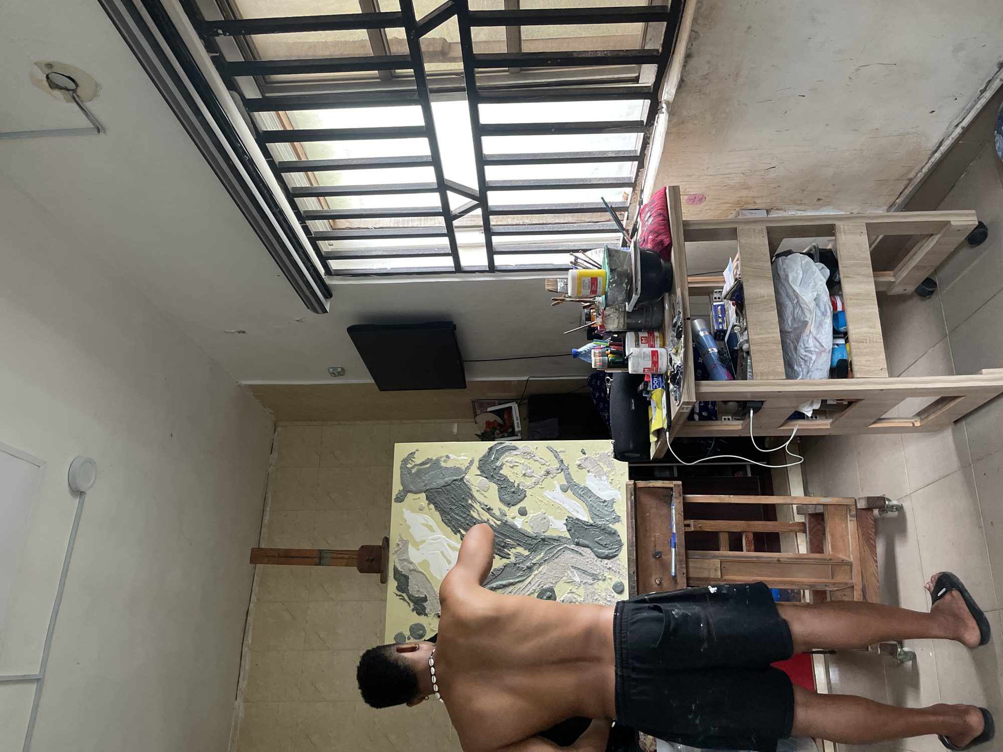 A man in black shorts and flip-flops stands in an art studio. He is facing an easel displaying an abstract painting in shades of gray and white on a yellow background. He is giving a thumbs-up. An art table with assorted paints and supplies is located behind him. The studio has a window with a metal grate and a ceiling fixture. The floor is tiled, and the walls are painted in neutral colors. The location is in Ado, Nigeria.