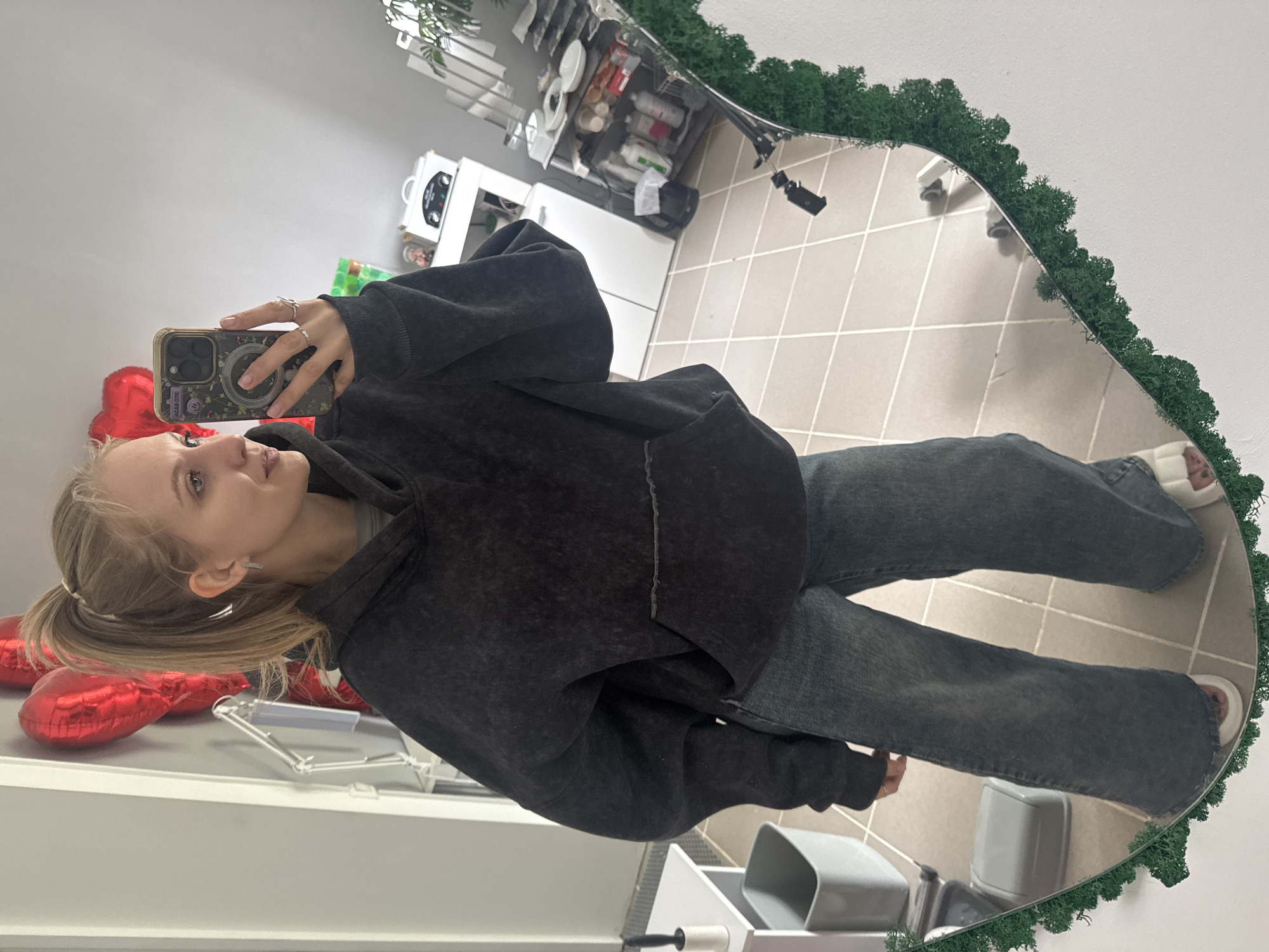 A woman with light blonde hair is taking a selfie in a mirror with a mossy green border. She is wearing a dark gray hooded sweatshirt and blue jeans. The mirror reflects a tiled floor and a shelf with various objects. To the left of the woman, red heart-shaped balloons are visible. The location is likely an interior space, but specific details indicating the city of Izhevsk, Russia, are absent.