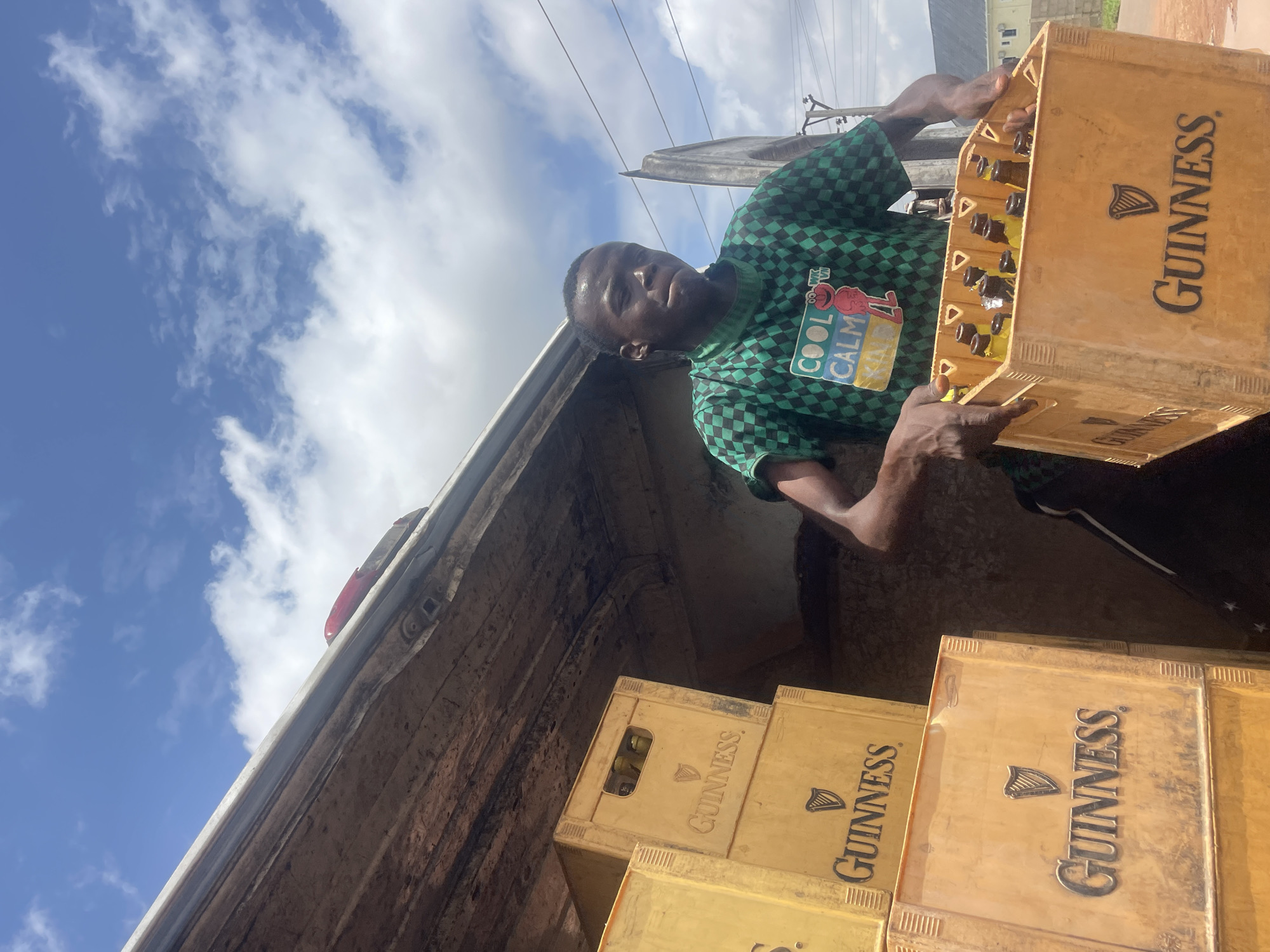 In Gura Top, Nigeria, a young man in a green checkered t-shirt is seen standing next to a truck, loading or unloading crates of Guinness. The crates are yellow plastic and stacked high. The man is holding one crate of Guinness bottles in his hands, which is filled with amber liquid and has dark caps. The truck bed is visible, made of weathered wood. The sky above is a bright blue with scattered white clouds. The surrounding environment appears to be an outdoor urban area, with a hint of buildings and utility poles in the background.