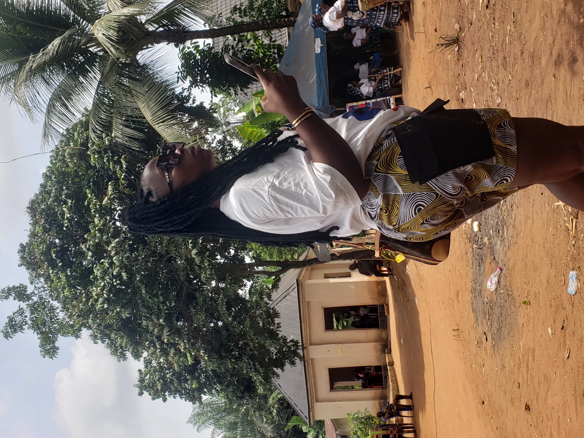 A person stands outdoors in Umundugba, Nigeria, holding a mobile phone. The individual wears a white t-shirt, patterned shorts, and sunglasses. The environment includes a building with window openings, trees including a palm tree, and bare ground littered with plastic bottles. Other individuals are visible in the background.