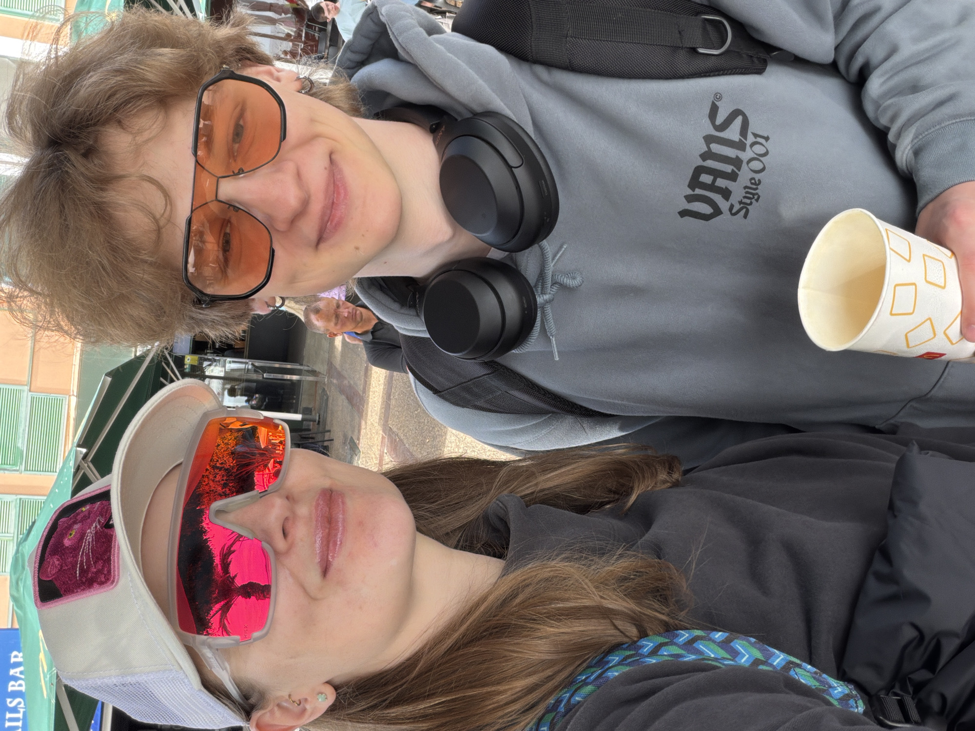 A selfie-style image, rotated 90 degrees counter-clockwise, features two young individuals smiling at the camera in what appears to be an outdoor public setting, possibly Los Cristianos, Spain.
The person positioned at the top, a male with light brown hair, wears large orange-tinted aviator-style sunglasses that reflect parts of the environment. He is dressed in a grey hoodie, which clearly displays the black "VANS" logo with "Style OO1" underneath it on the chest. Black headphones rest around his neck, and a black backpack strap is visible over his right shoulder. He holds a white paper cup patterned with orange squares in his right hand.
Below him, a female with long brown hair also smiles at the camera. She wears striking mirrored pink and orange sunglasses, which show reflections of what appears to be trees or palm fronds. Her head is adorned with a white trucker hat featuring a mesh back and a distinctive pink panther patch on the front. She is wearing a dark grey or black jacket, and a blue and green patterned strap is visible over her shoulder. A small ear piercing is visible on her left ear.
The background is somewhat blurred but shows glimpses of buildings, green structures like awnings or blinds, and other indistinct figures, indicating a bustling public space. A blue sign with partial text "AILS BAR" is visible behind the female subject. The bright lighting suggests it is daytime, and the presence of sunglasses on both individuals implies sunny weather. Both subjects exhibit cheerful expressions.