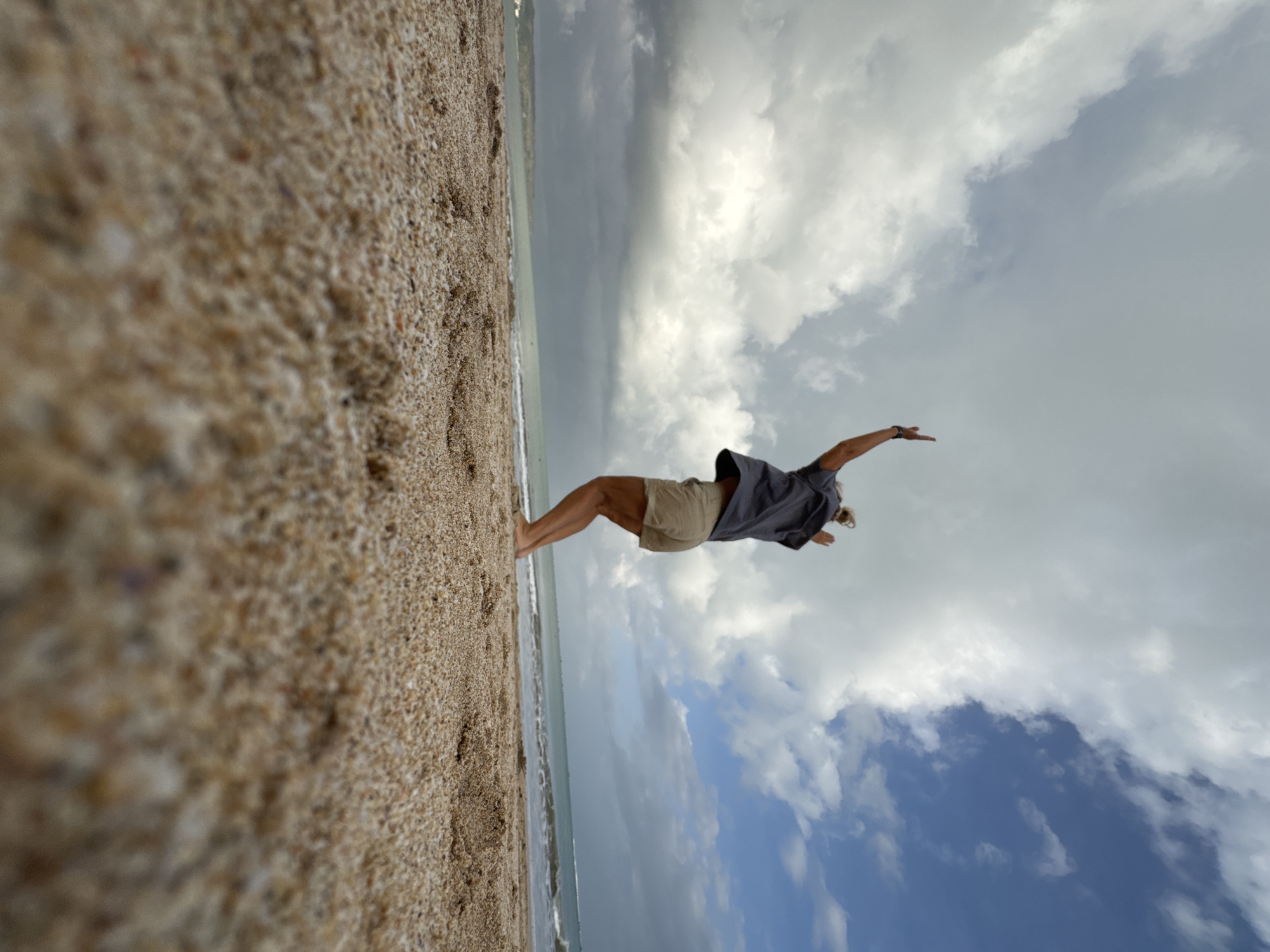 A low-angle, wide-shot photograph captures a person jumping on the sandy shore of Kuta Beach, Indonesia. The individual, viewed from behind, is mid-air with arms raised, wearing a dark t-shirt and tan shorts. The foreground is dominated by coarse, light-brown sand, sharply in focus and filling the bottom half of the frame. Beyond the sand, the ocean stretches out, its turquoise water meeting a distant, hazy coastline. The sky above is a dramatic mix of dark, heavy clouds on the left and bright blue sky with scattered white clouds on the right, suggesting rapidly changing weather. The waves are gentle, lapping at the shore. The overall impression is one of movement and freedom against a dynamic natural backdrop.