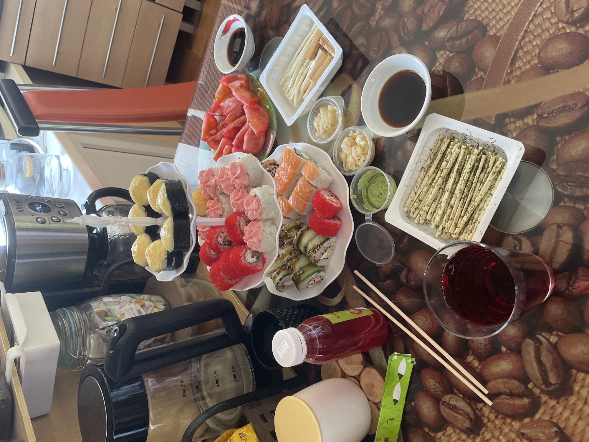 The image shows a table laden with various dishes of sushi, including rolls with red, pink, and orange toppings. There are also sides such as sliced tomatoes, cheese sticks, ginger, and wasabi. Liquids are present in bowls and a glass. Chopsticks are also visible. In the background, kitchen appliances like a coffee maker are noticeable, along with a glimpse of orange seating. The table has a patterned surface resembling coffee beans. Based on the presence of a bottle labeled in Russian, the location is likely Russia, and given the specific style of the scene, it may be in Perm, Russia.