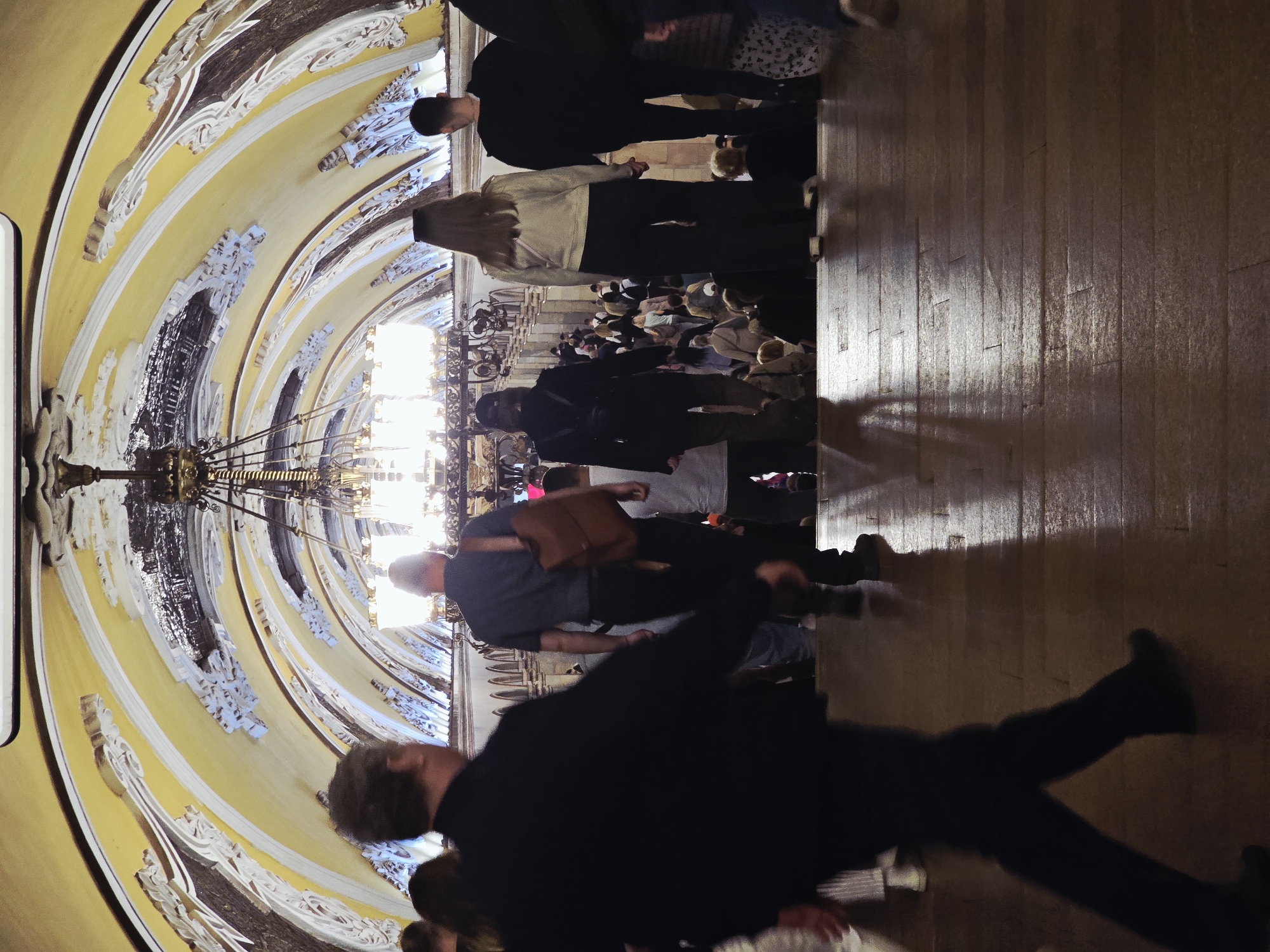 The image shows a hall or corridor in what appears to be a public building, possibly a metro station or a museum, in Moscow, Russia. The architecture is ornate with elaborate plasterwork and paintings on the ceiling, and a large chandelier hangs in the center. People are walking through the space; some are in the foreground and appear blurred due to movement, while others are in the background and more in focus. The floor is made of wood panels. The overall impression is of a grand and busy public space.
