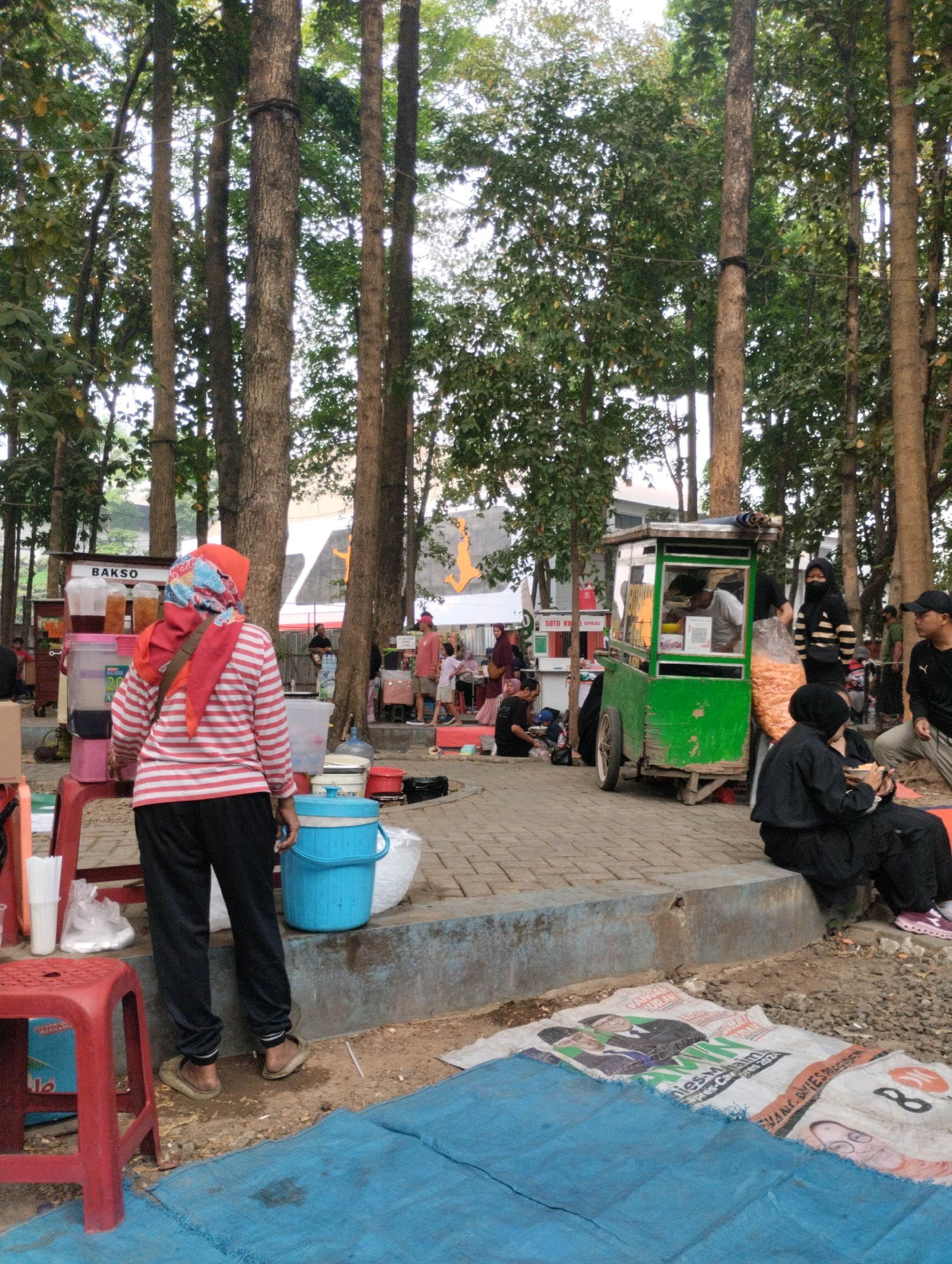 The image depicts an outdoor scene with numerous tall trees and a bustling market area. In the foreground, a person wearing a red and white striped shirt, black pants, and a red headscarf stands facing away from the camera. A red plastic stool is to the left, and a blue cooler is in front of the person. Behind the cooler, there are various items, including plastic bags and a large blue tarp spread on the ground.
To the right of the foreground person, a green food cart is visible, with people attending to customers and preparing food. A woman in a black hijab and striped shirt stands next to the cart, holding a clear plastic bag filled with snacks. Other individuals are seated and standing around the food cart. Further in the background, several more food stalls are set up, indicated by the presence of tables, chairs, and various goods for sale.
The overall setting suggests a lively outdoor market or food court, likely in a public space in Bekasi, Indonesia, given the presence of food vendors and the general atmosphere. The ground is a mix of paved walkways and dirt. Sunlight filters through the trees, casting shadows and illuminating parts of the scene.