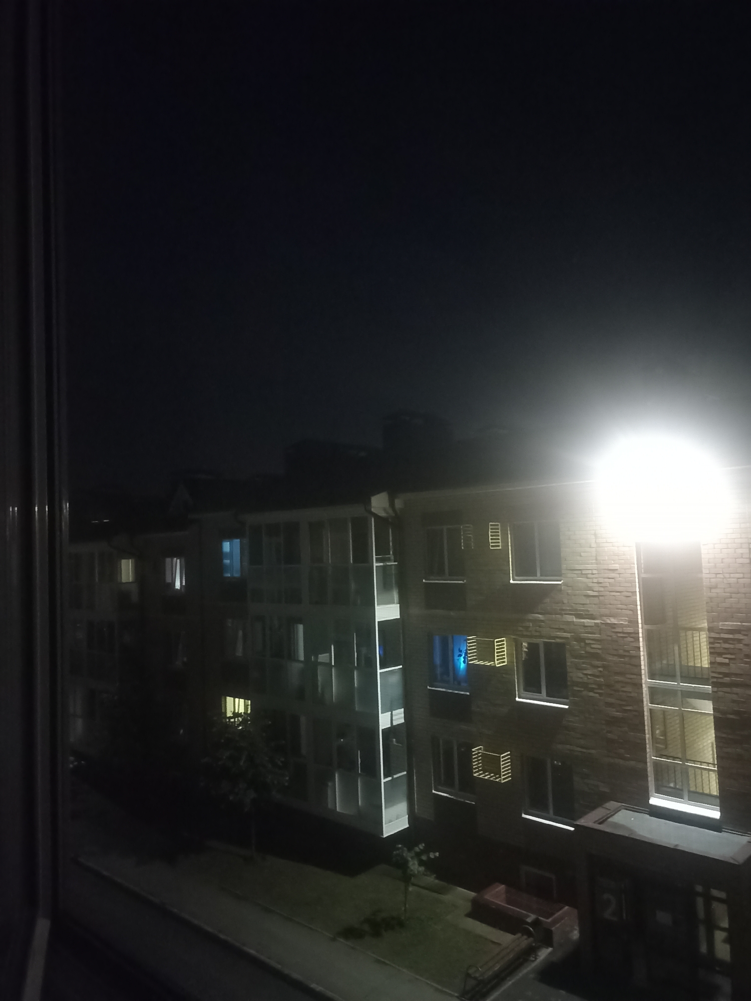 The image depicts a night scene in the city of Novoye Shigaleevo, Russia. A multi-story apartment building is visible, constructed with a combination of brick and white-paneled sections. Several windows are illuminated, with one on the third floor of the brick section emitting a distinctive blue light. A bright street lamp or floodlight illuminates the upper portion of the building and the sky. In the foreground, through a window frame, a dark path or sidewalk runs in front of the building, flanked by greenery. There are no people visible in the frame.