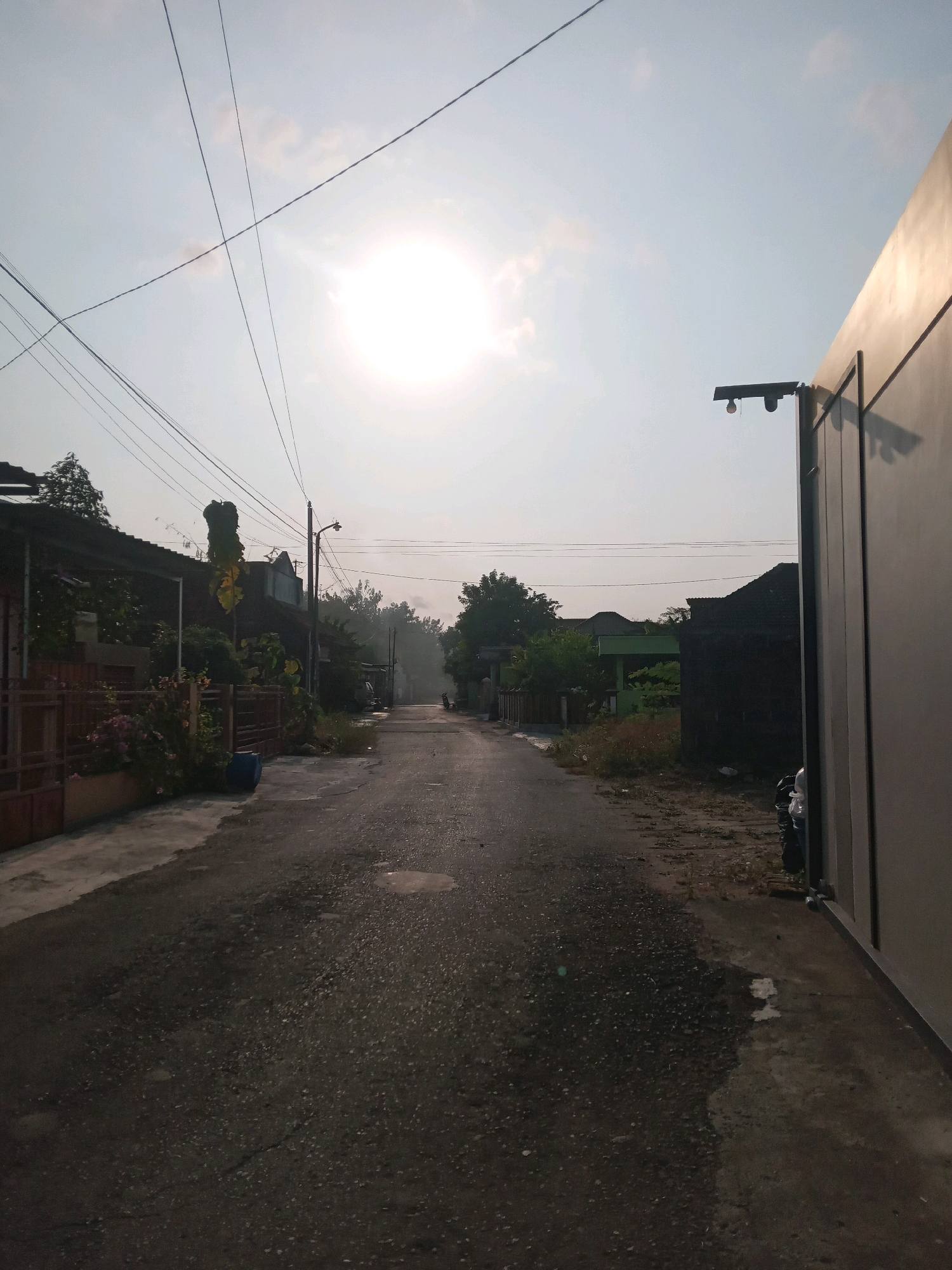 The image depicts a straight, asphalt road in the village of Sitimulyo, Indonesia, at sunrise. The sun, a bright orb, is positioned high in the sky, casting a soft glow and creating a hazy atmosphere. Overhead power lines crisscross the frame, adding to the rural, residential feel of the scene. On either side of the road are houses with dark roofs, some with fences and vegetation. A single lamppost stands near the middle of the road, suggesting it is a residential street. The road surface is wet, with several puddles reflecting the sky. To the right of the frame, a tall, smooth, beige-colored structure dominates the foreground, with a security camera mounted on its upper edge. A parked motorcycle is visible behind the structure, partially obscured. The overall impression is of a quiet, early morning in a suburban Indonesian village.