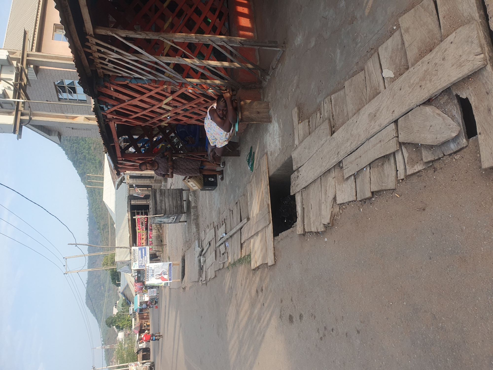 The image shows a street scene in Lokoja, Nigeria. The shot appears to be taken in an area with informal businesses or shops alongside the road. The road is paved but shows wear and tear. A drainage ditch runs alongside the road and is partially covered with wooden planks. A shop is on the right with a red metal frame. Two people stand and sit near the shop. One woman is sitting with her head resting in her hands, while the second is standing. In the background, other buildings, hills, and power lines are visible. There are also signs and advertisements further down the street. A few individuals can be seen riding bicycles further down the road.