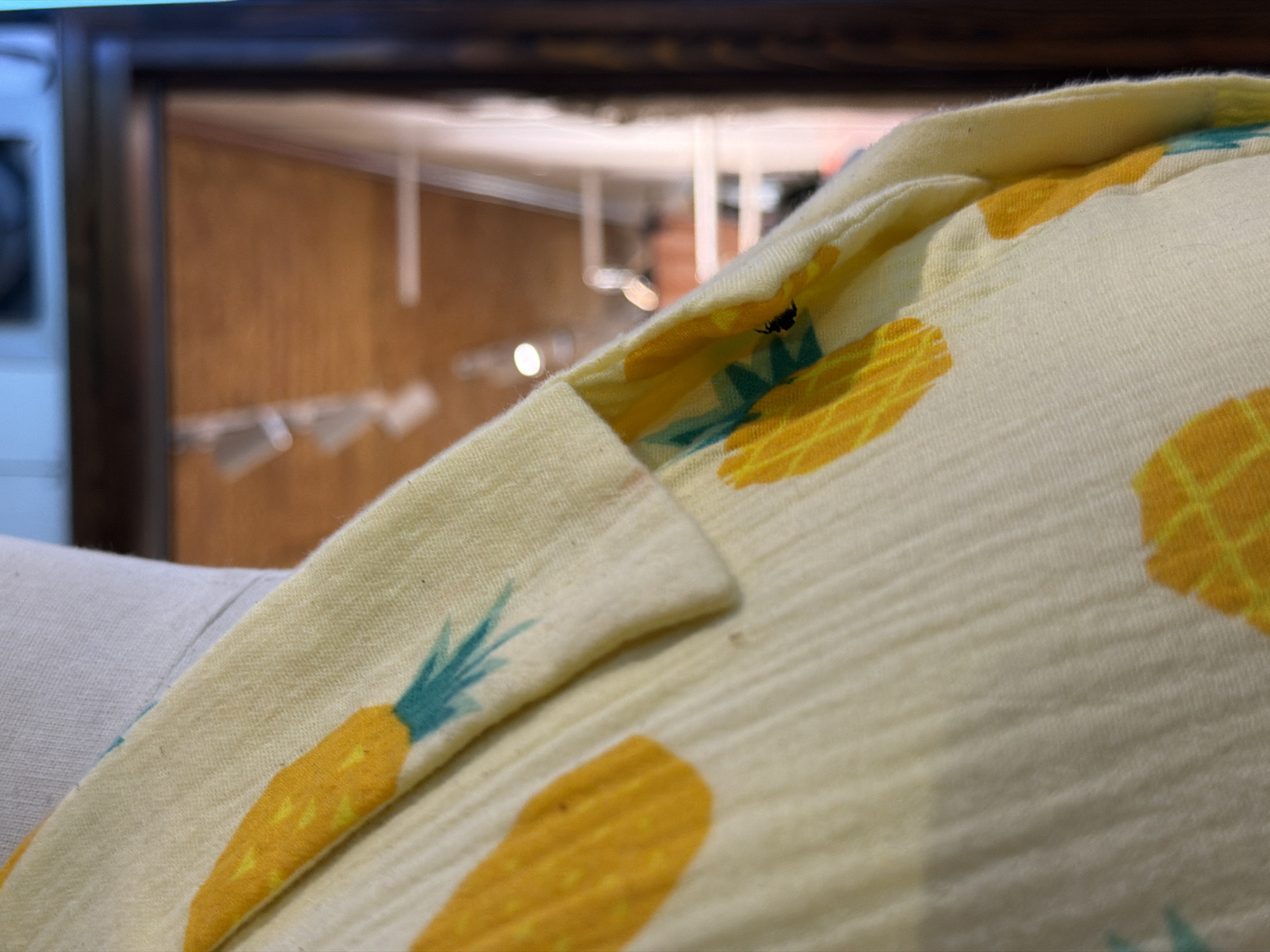 A close-up shot shows a piece of clothing with a pineapple pattern. The fabric is light yellow and has a textured surface. Each pineapple is yellow with a blue-green top. An insect, possibly an ant, is visible on the fabric. The background is blurry but shows a reflection of an interior space with a wooden frame.
There is no information in the image to confirm if the location is in Đà Nẵng, Vietnam.