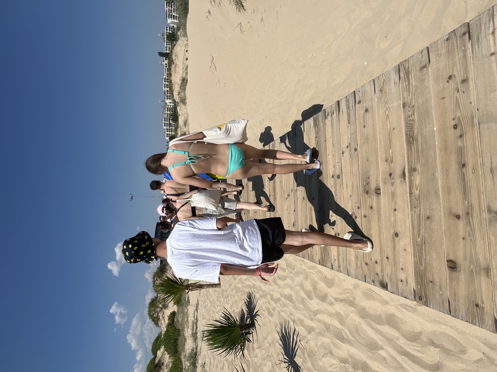 A group of people are walking on a wooden boardwalk through sandy dunes towards buildings in Nesebar, Bulgaria. The boardwalk is made of weathered wooden planks and is bordered by sand. Low-lying green bushes and palm-like plants are visible in the dunes to the left. The sky is a clear blue with a few scattered white clouds. In the distance, several modern white buildings are visible, suggesting a resort or residential area.
The person in the foreground is a young male wearing a white t-shirt, black shorts, and a black bucket hat with a yellow pattern. He is wearing white sandals. A red wristband is visible on his left wrist. Behind him, a young female is walking in a turquoise bikini, carrying a white tote bag. She is wearing dark flip-flops. Other people are visible further down the boardwalk, some carrying bags. The overall scene suggests a sunny day at a beachside location.