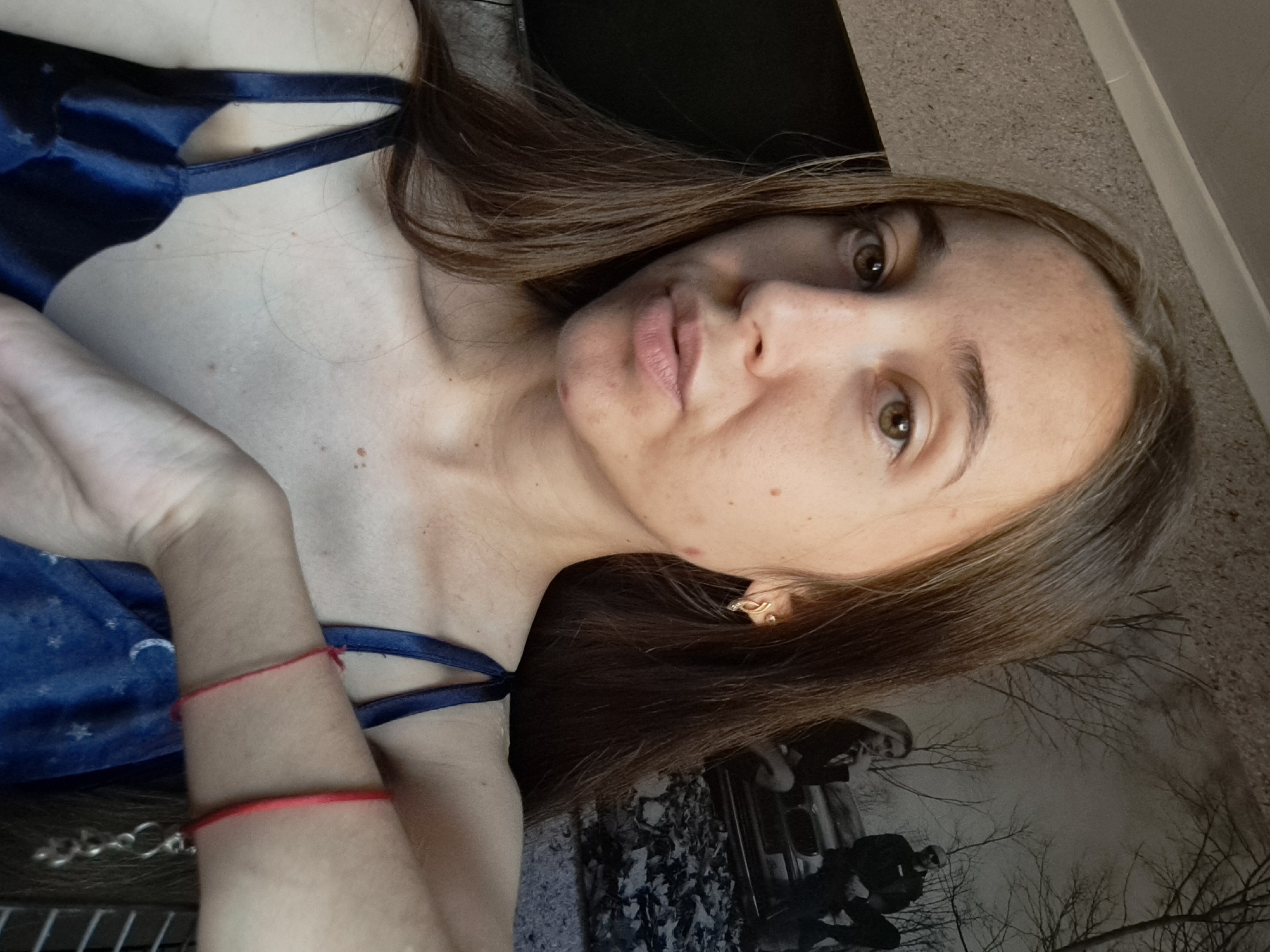 The image shows a young woman with long, brown hair, looking directly at the camera. She is wearing a dark blue satin slip with thin straps, adorned with a crescent moon and star pattern. A thin red bracelet is visible on her right wrist. Her skin has some visible blemishes and freckles. She is in an indoor setting, with a textured wall and a large black and white print on the wall behind her, depicting trees and two figures sitting on what appears to be a vehicle. The overall lighting suggests a daytime setting. The image does not contain any specific details to indicate its location within the city of Vypasne, Ukraine.