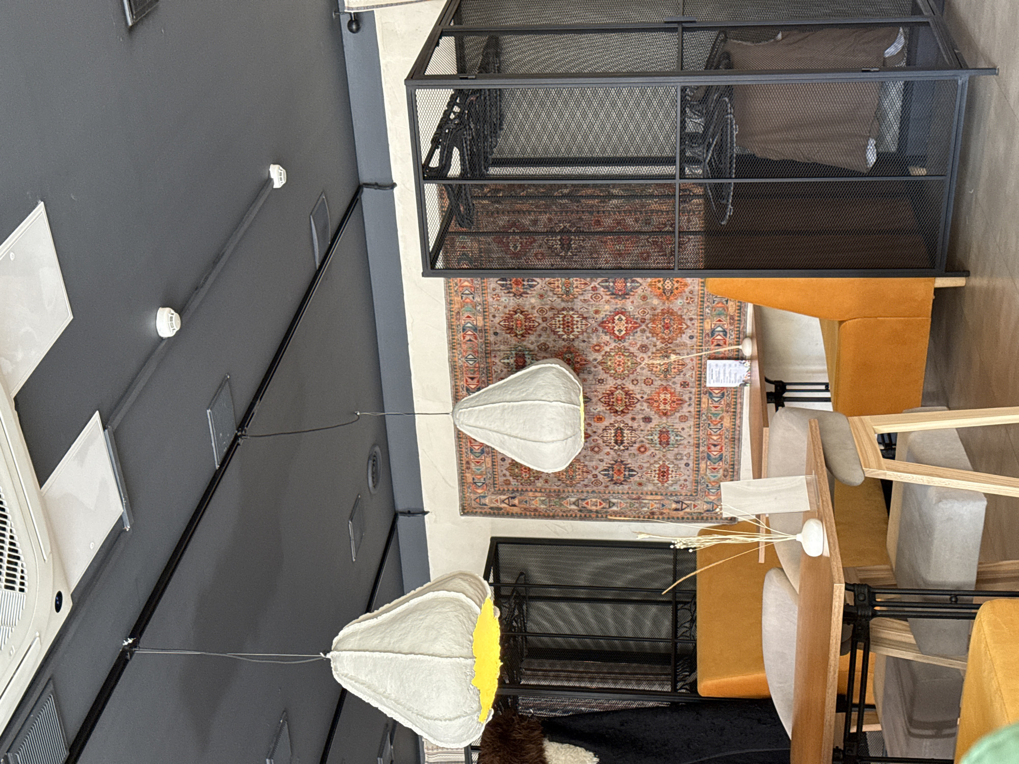 This media file captures an indoor commercial space, likely a furniture showroom, cafe, or retail display area, viewed from an upward angle. No people are visible in the scene.
The ceiling is dark grey or black, featuring several square and circular HVAC vents, white circular lights (possibly smoke detectors or recessed fixtures), and a black conduit running across it. Two distinctive, lantern-like light fixtures hang from the ceiling at different depths. These fixtures are crafted from a textured, light-colored material, resembling pleated paper or fabric, with one revealing a bright yellow interior.
The floor is covered by a large, ornate oriental rug displaying intricate patterns in shades of red, orange, blue, and beige. Positioned on the rug are various furniture pieces and display elements. A prominent black metal mesh unit, possibly a shelving system or room divider, stands tall, containing what appears to be textiles or cushions. In front of and around this unit, several upholstered benches or armless sofas in a vivid mustard yellow color are arranged. Nearby, several wooden chairs with light grey upholstered seats and backs are visible, some appearing stacked.
On a surface, likely a table or counter, a small white vase holds dried decorative reeds or grasses. A white sign or piece of paper with text is also visible on a surface near a yellow sofa; the word "INFORMA" can be partially discerned at the top.
The overall impression is that of a modern interior design with an industrial edge, softened by traditional elements and warm-toned furniture. The space is well-lit by artificial light, making the time of day indeterminate. There are no specific activities taking place, and the scene appears static, focused on showcasing the decor and furnishings.