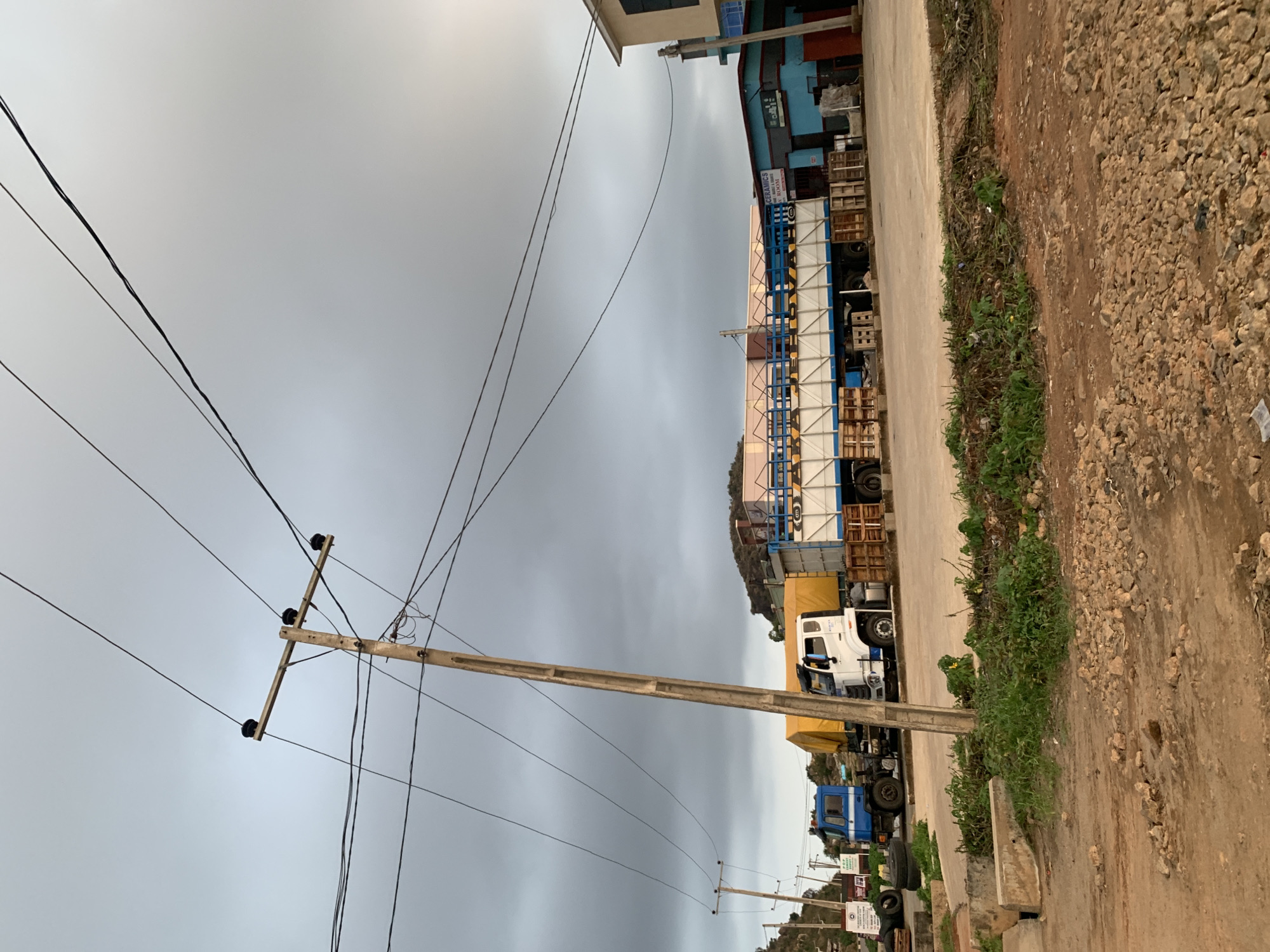 The scene is Gura Top, Nigeria, presenting a view of a roadside. The sky is overcast. A line of utility poles runs along the left side of the image. Power lines are strung between the poles. Along the right edge is a dirt road and some vegetation. Parked along the paved road is a line of trucks, some carrying cargo, possibly building materials. The first truck in the line is blue, followed by a white truck, and then one with a yellow tarp. A building is partially visible in the upper right corner.