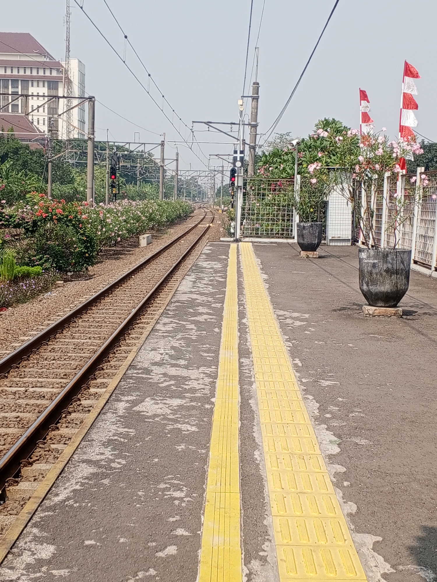 The image depicts a train station platform in Depok, Indonesia, with two sets of train tracks. The platform is paved with asphalt and features a raised yellow tactile paving strip along the edge of the tracks. On the left side of the image, a lush row of flowering bushes with red and pink blooms lines the train tracks. To the right, a white metal fence encloses a small garden area with more flowering bushes and two large dark gray planters. One planter has the word "Commuter" written on it. Several Indonesian flags, red and white, are visible flying from poles behind the fence. In the background, a tall building with a white facade and a brown roof stands against a pale sky, along with utility poles and overhead electrical wires for the trains. A red traffic signal is visible on a pole next to the tracks. The scene is set during daylight, with clear weather.