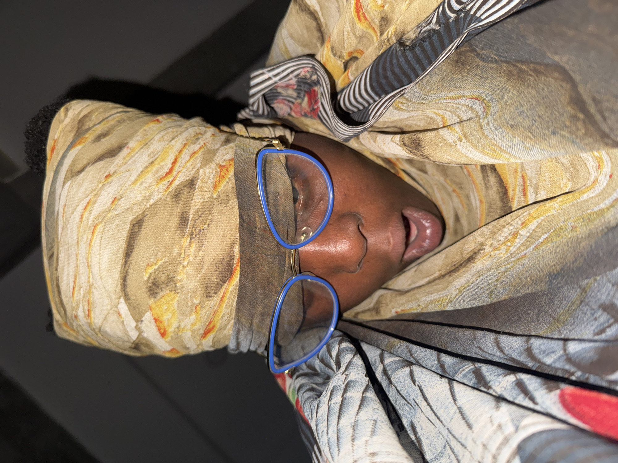 The image shows a close-up, low-angle shot of a person wearing glasses and a patterned headscarf. The person's mouth is open, and their eyes are closed. The headscarf is a light brown color with swirling patterns of yellow, orange, and white. The person is also wearing a shawl or scarf that drapes around their shoulders, featuring a pattern of gray, white, and black stripes and a floral design with red accents. Blue-rimmed glasses with gold arms are worn on the face. The background is dark and out of focus, making it impossible to determine the environment. There is no information available about the location in Yelwa, Nigeria.