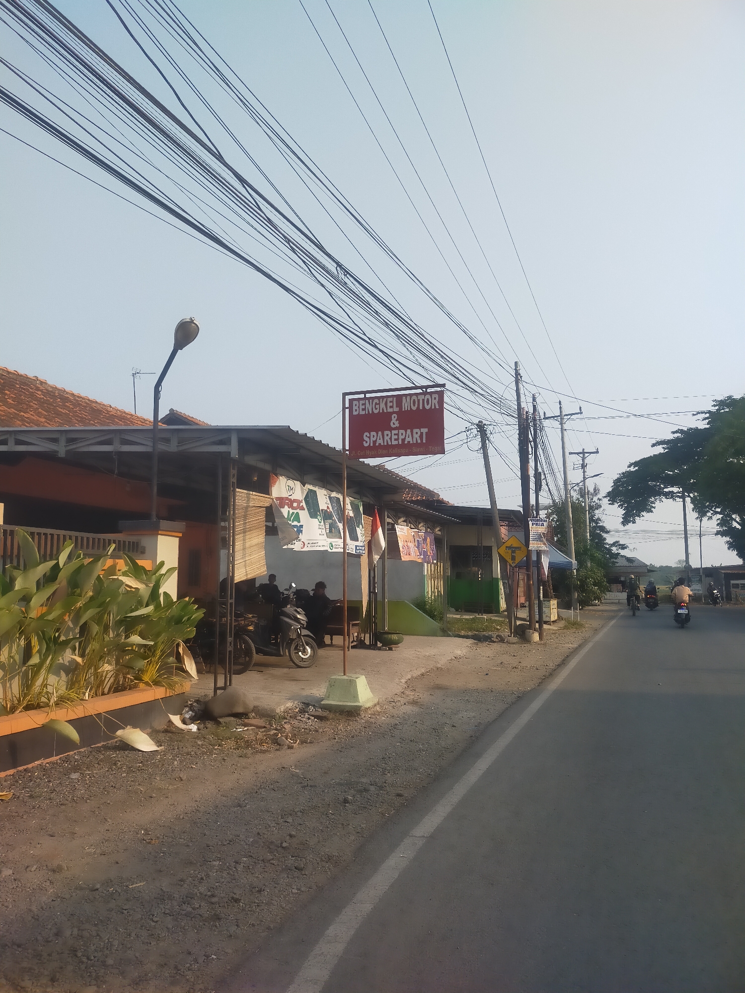 The image depicts a roadside view in Slawi, Indonesia, showcasing a "Bengkel Motor & Sparepart" (Motorcycle Workshop & Sparepart) establishment. The workshop is a low-rise building with a corrugated metal roof supported by metal pillars. In front of the workshop, several motorcycles are parked, and two individuals are seated, appearing to be engaged in conversation or work. A red sign with white lettering announces the workshop's name. Adjacent to the workshop, a street lamp stands tall, and numerous electrical wires crisscross the sky overhead. The street itself is paved and appears to be a two-lane road, with a few motorcycles in motion further down. The roadside is lined with gravel and dirt, with some vegetation and debris present. The overall scene suggests a typical commercial area in a provincial Indonesian town, likely on a clear, sunny day given the lighting.