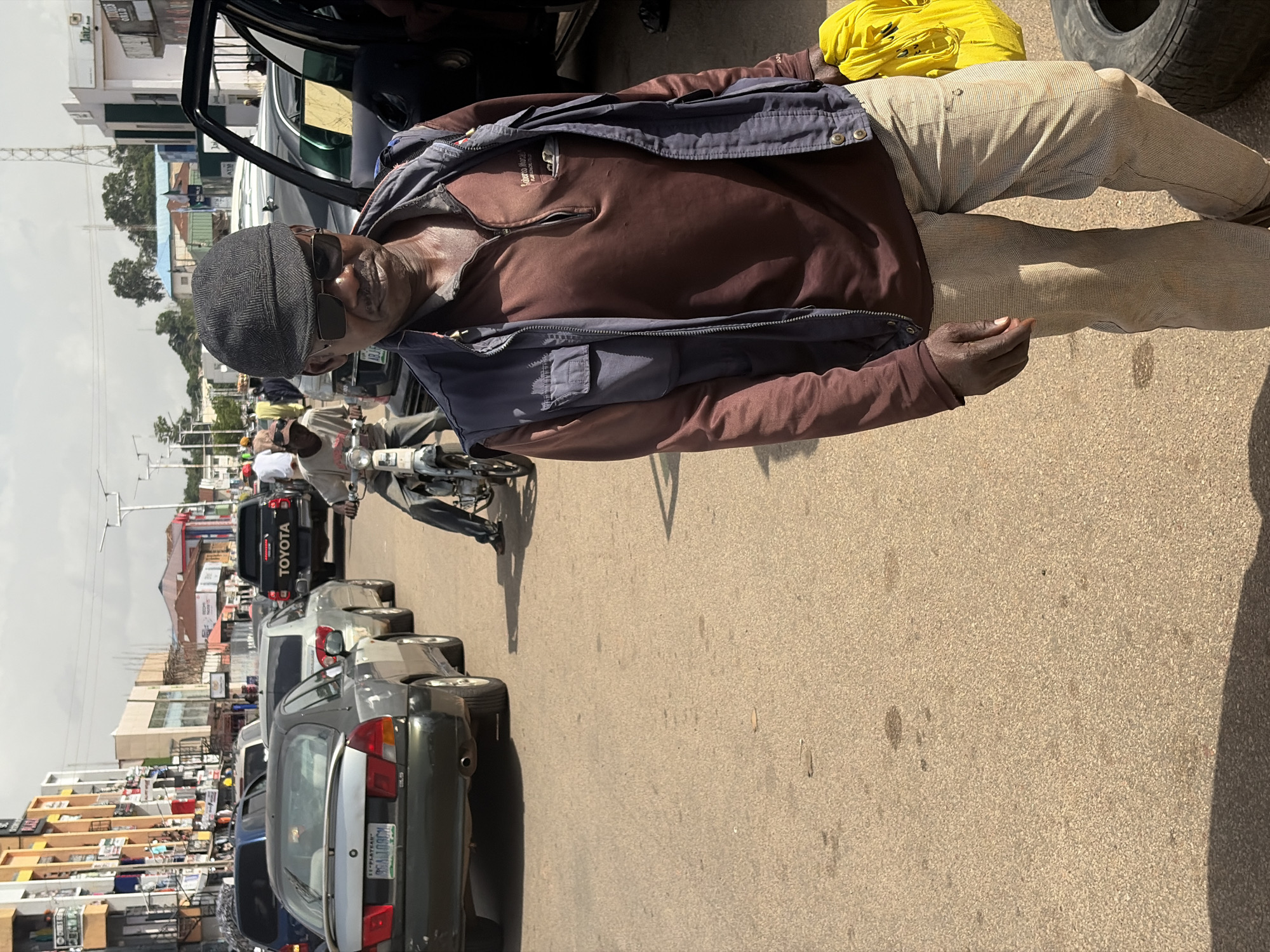 The image depicts a street scene in Jos, Nigeria. A man in the foreground, wearing a cap, sunglasses, a maroon shirt, and a dark vest, stands on a paved road. He is holding a bright yellow bag in his right hand. Behind him, traffic is present, with cars and a motorcycle visible. A black Toyota pickup truck is in the middle ground. Buildings line the street on both sides, with some showing commercial signage. The sky is overcast. The environment appears to be an urban setting with moderate traffic congestion.