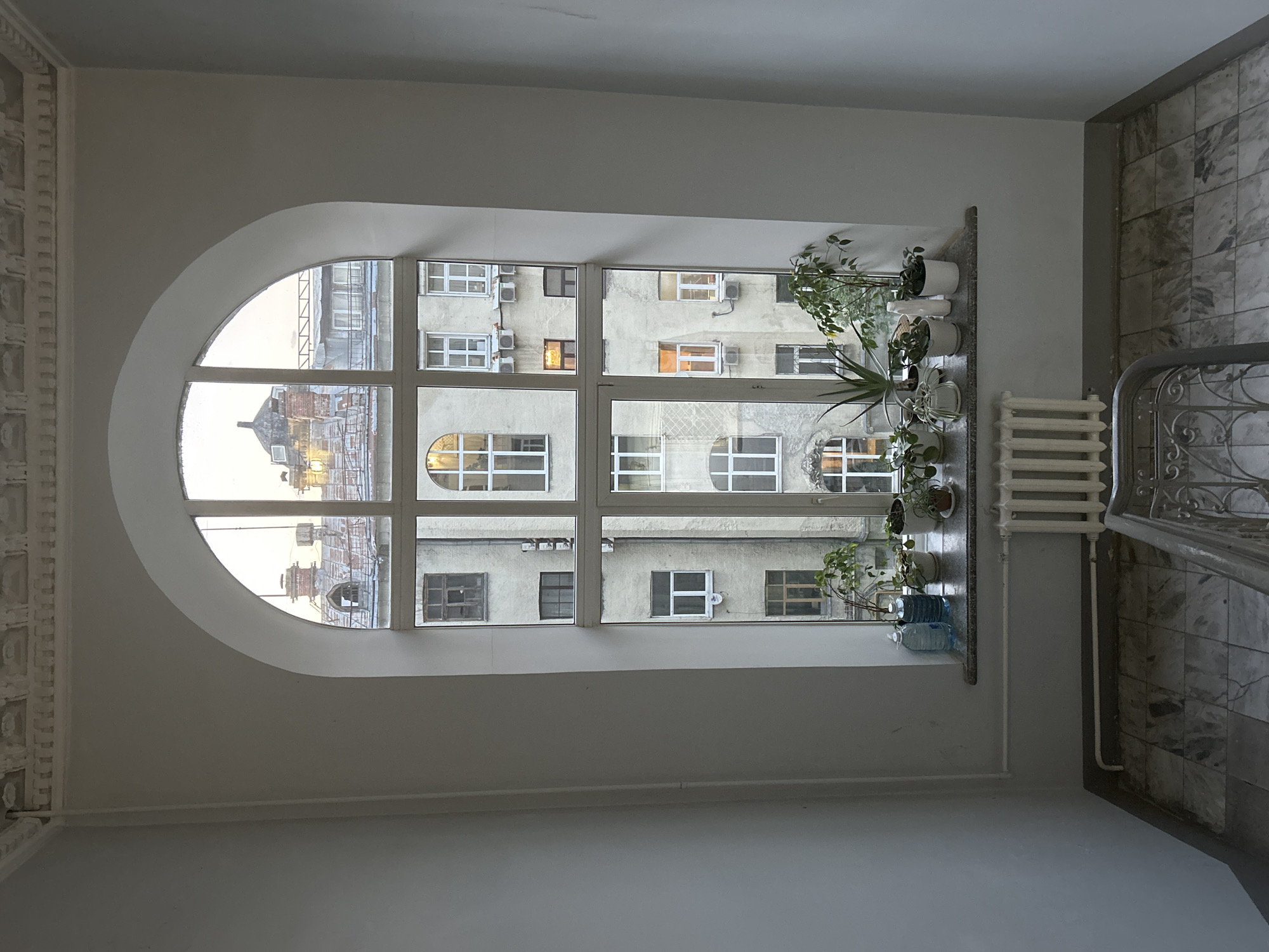The image captures an interior space with a prominent arched window looking out onto a building across an alley. The window is divided into multiple panes and displays the exterior of an aged building with numerous windows, some containing air conditioning units. On the window sill, a collection of potted plants is arranged. A radiator is positioned to the right of the window, adjacent to a marble staircase. The walls are painted white, contributing to a neutral backdrop. The location appears to be in an urban environment, although the exact city is undetermined. The scene is static and depicts an intersection of architectural elements, natural light, and domestic objects.