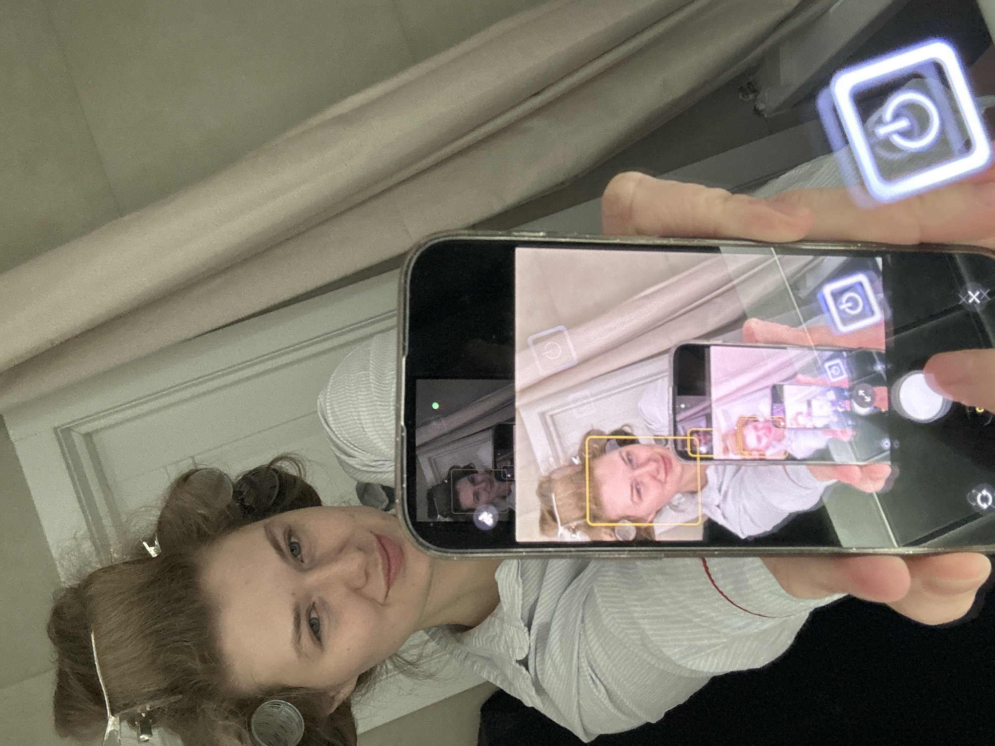 A woman is photographing herself in a mirror using a smartphone. The reflection in the mirror shows a woman with brown hair and hair rollers. The woman is wearing a striped shirt. She is holding a smartphone in her right hand and pressing the capture button. The smartphone screen is displaying the woman taking the photo. The image includes an augmented reality (AR) element of a glowing power button. The location appears to be a residential interior, likely a bathroom, in Ключищи, Russia.