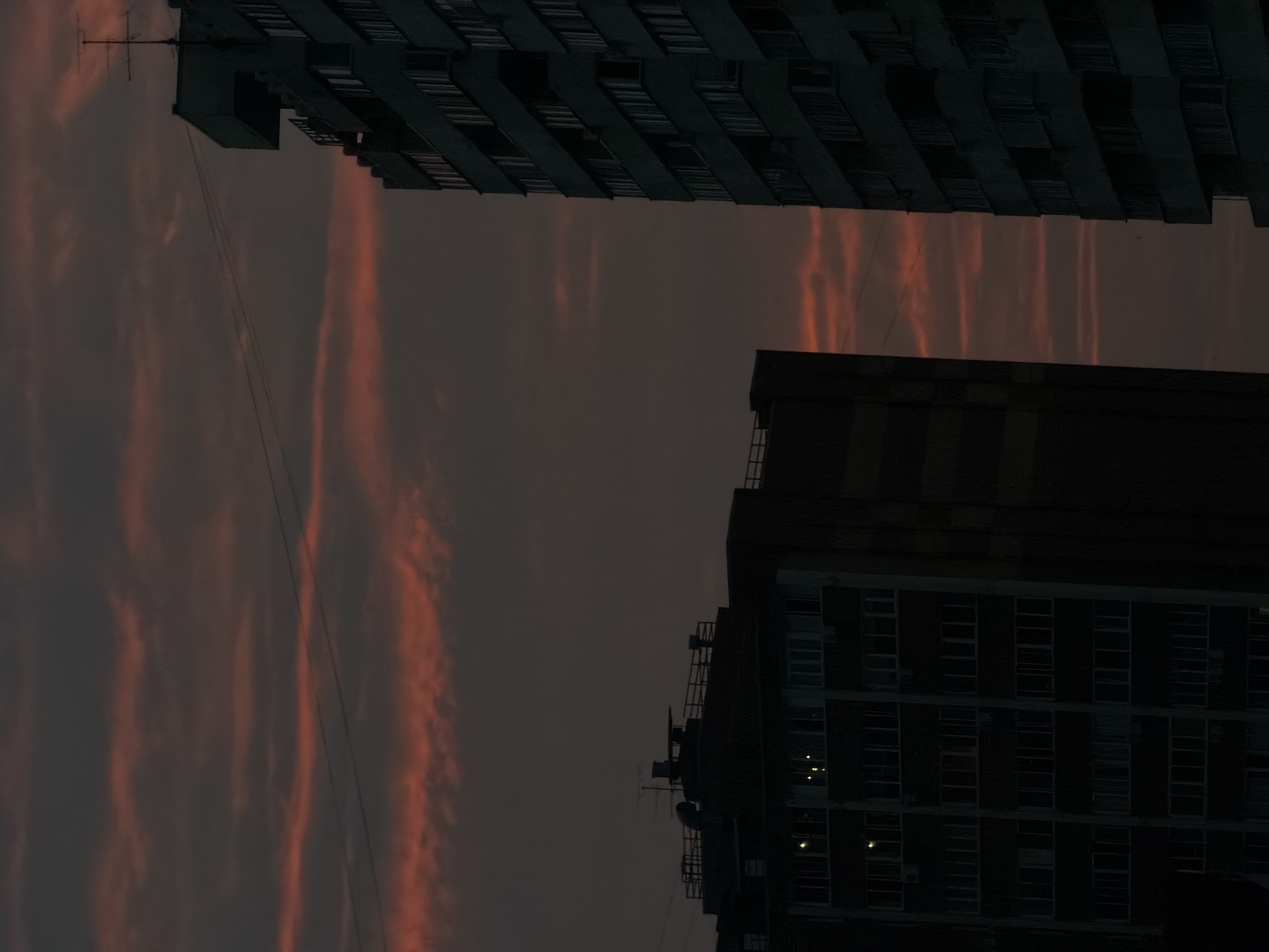 This media file shows a low-angle view of two dark, silhouetted apartment buildings against a dusky sky. The sky is filled with wispy, orange-red clouds, suggesting a sunset or sunrise. Power lines are visible, stretching between the buildings and across the sky. The buildings appear to be constructed of concrete and have rows of dark windows. Some windows show faint yellow lights, indicating occupancy within the apartments. The overall mood is somber and urban, with the vibrant sky providing a dramatic contrast to the austere architecture. The image was taken in Novosibirsk, Russia. There is no visible text in the media file.