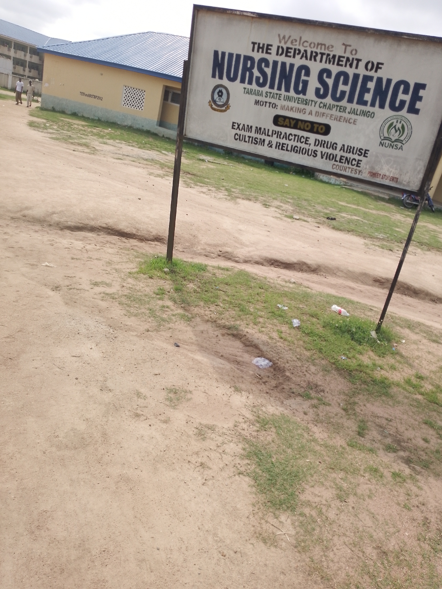 A close-up, slightly tilted outdoor shot captures a dusty, dirt path leading to a building. A signpost stands in the foreground, its metal pole rusted and planted in a patch of grass with scattered debris. The sign itself is rectangular with a white background, featuring bold, dark text that reads "Welcome To THE DEPARTMENT OF NURSING SCIENCE." Below this, smaller text specifies "TARABA STATE UNIVERSITY CHAPTER JALINGO" and "MOTTO: MAKING A DIFFERENCE." A yellow button with "SAY NO TO" is prominently displayed, followed by a list of prohibitions: "EXAM MALPRACTICE, DRUG ABUSE CULTISM & RELIGIOUS VIOLENCE." The sign also includes the acronym "NUNSA" and two circular emblems. In the background, a light yellow building with a blue roof is visible, with two individuals walking near its entrance. The ground is predominantly dry, sandy earth with sparse patches of green grass. The sky above is overcast.